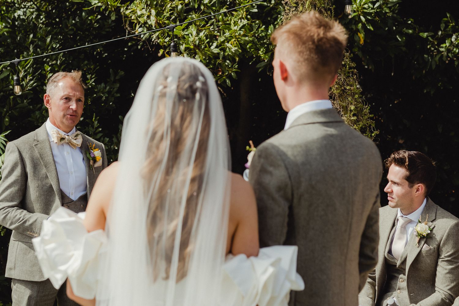Wedding party stands outdoors in formal attire during summer ceremony with lush greenery in background.