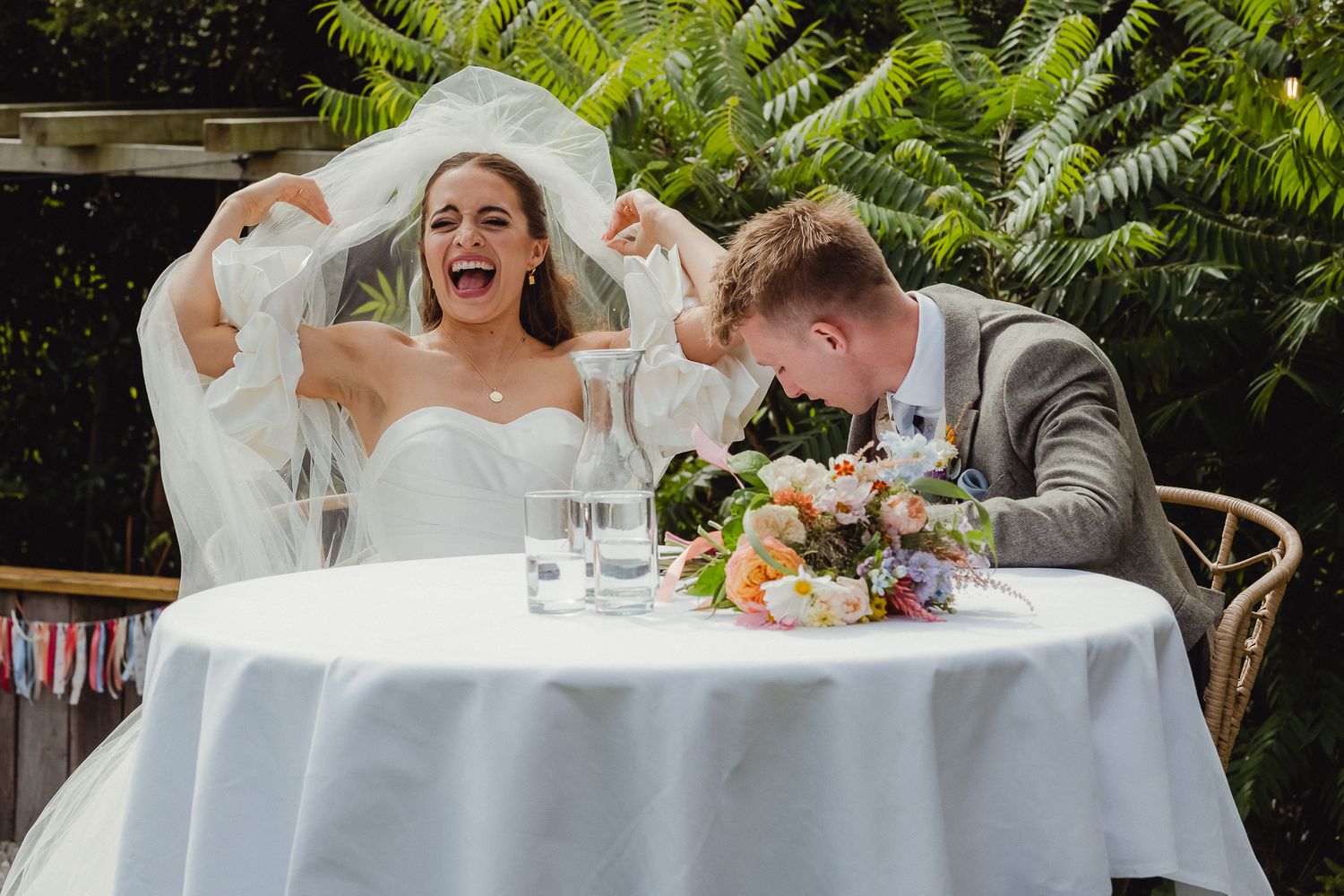 A wedding couple laughs joyfully while seated at an outdoor reception table surrounded by lush greenery.