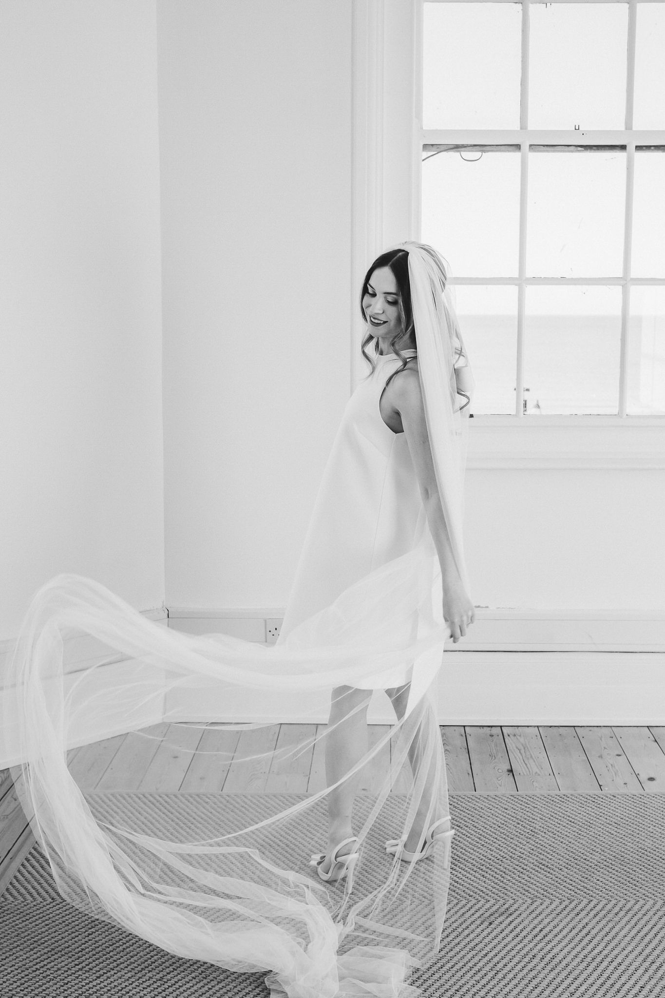 Black and white photo of a bride in a flowing veil twirling gracefully by a bright window in an elegant room.