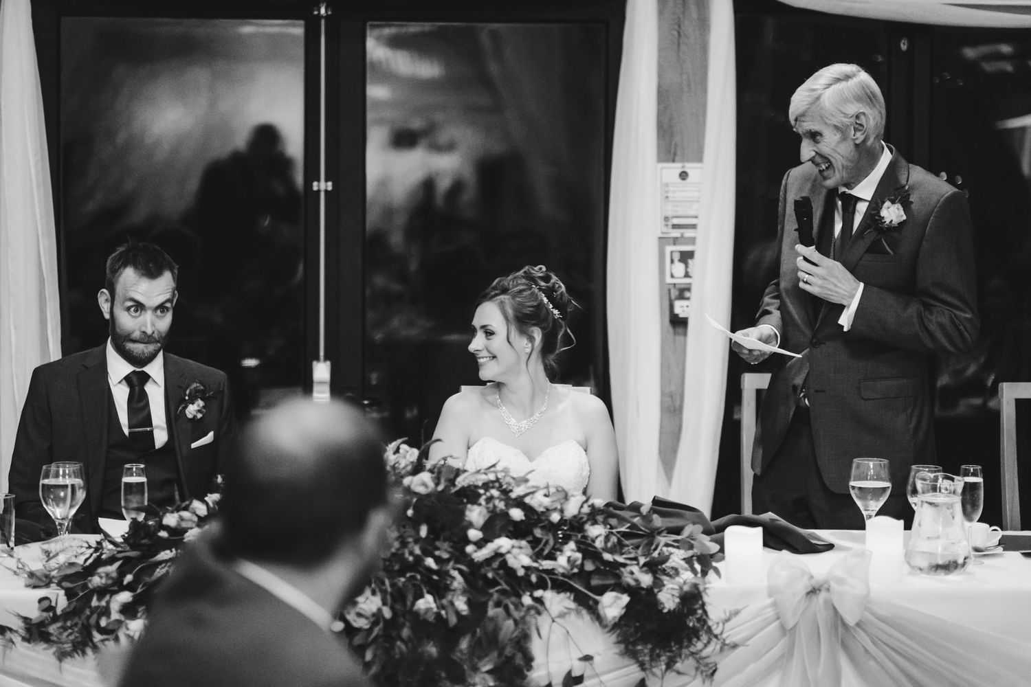 Black and white photo of wedding reception speeches at decorated head table.