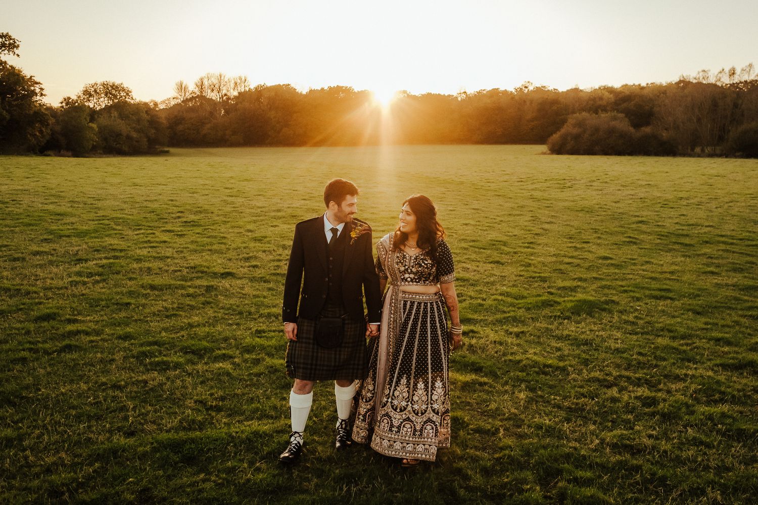 Fusion wedding couple in Scottish kilt and Indian dress standing together in golden field.