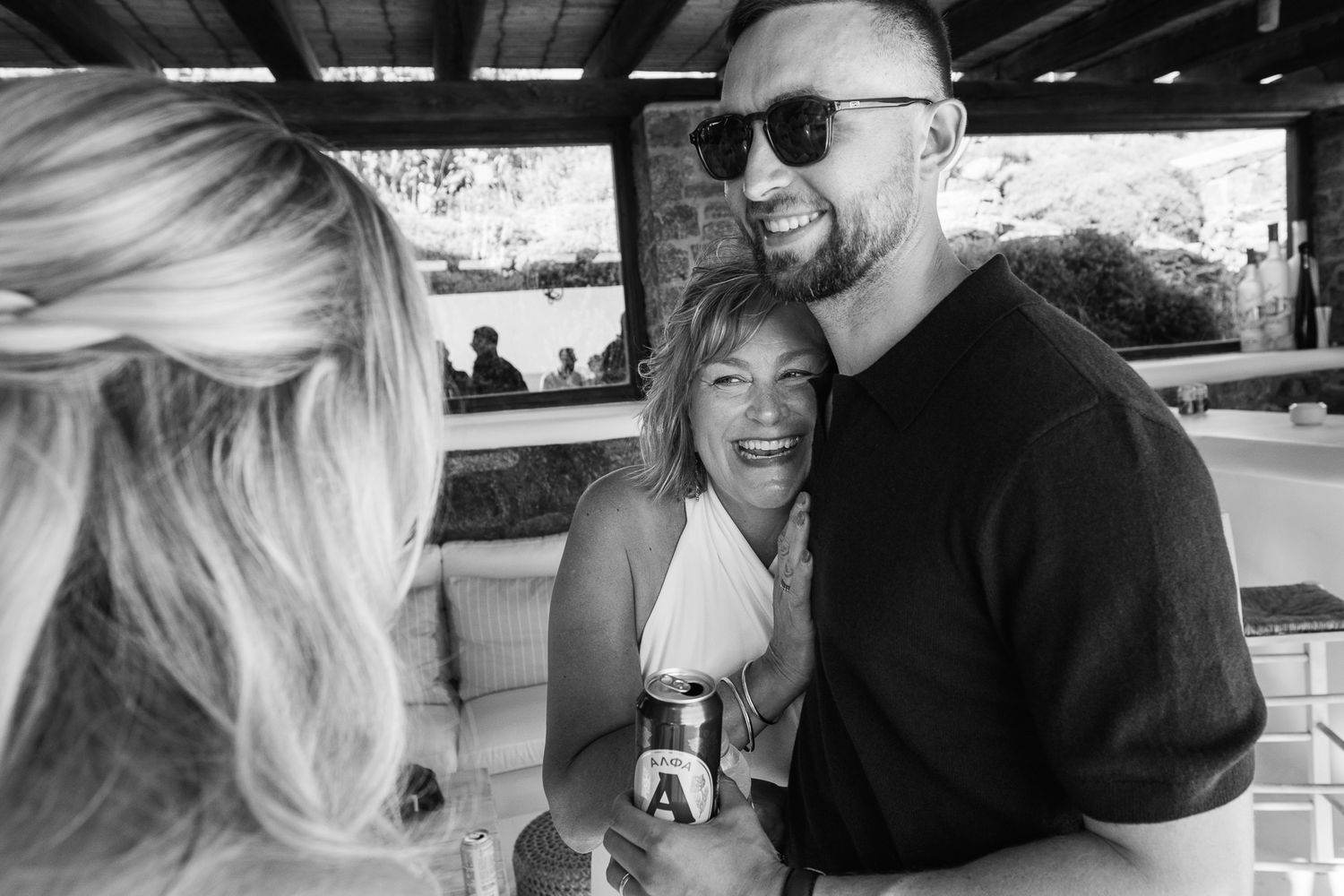 Black and white photo of a man in sunglasses hugging a smiling woman at an outdoor social gathering.