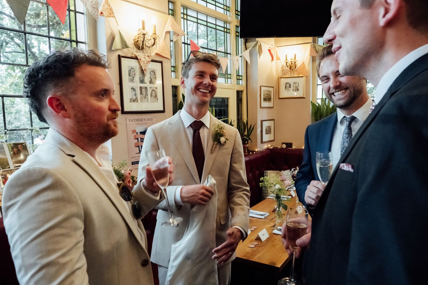 Three men in suits hold champagne glasses and chat at a wedding reception inside an elegant venue with framed art and large windows.