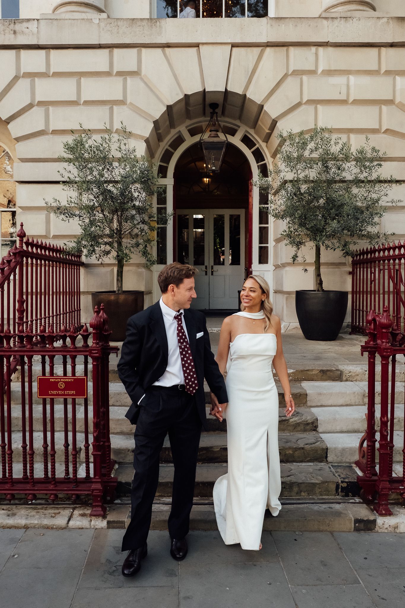 Bride in a white satin gown and groom in a dark suit hold hands on stone steps before an arched doorway with red iron railings.