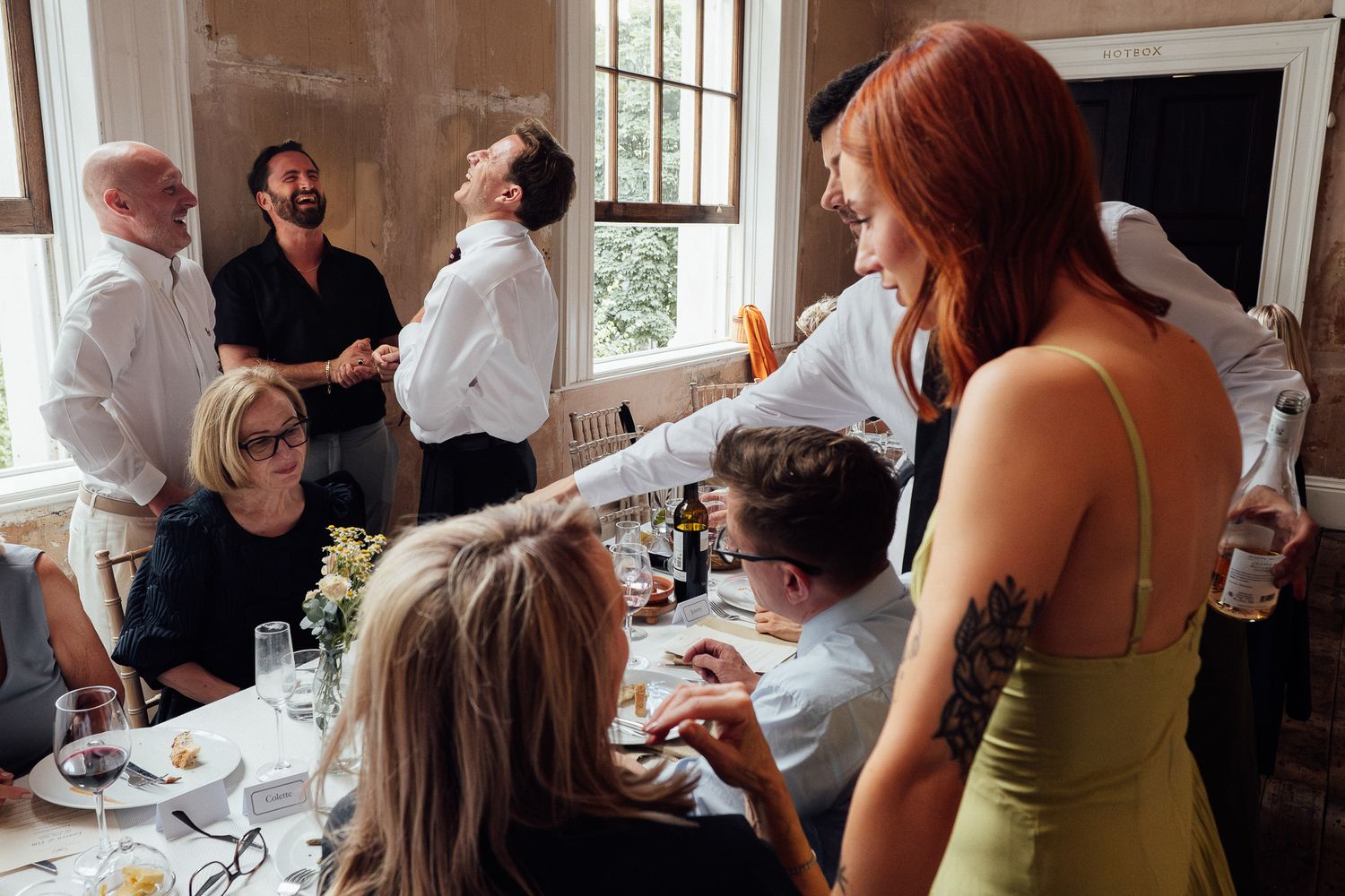 Guests laugh and chat around a dining table at a lively dinner party, with a red-haired woman in a green dress standing in the foreground.