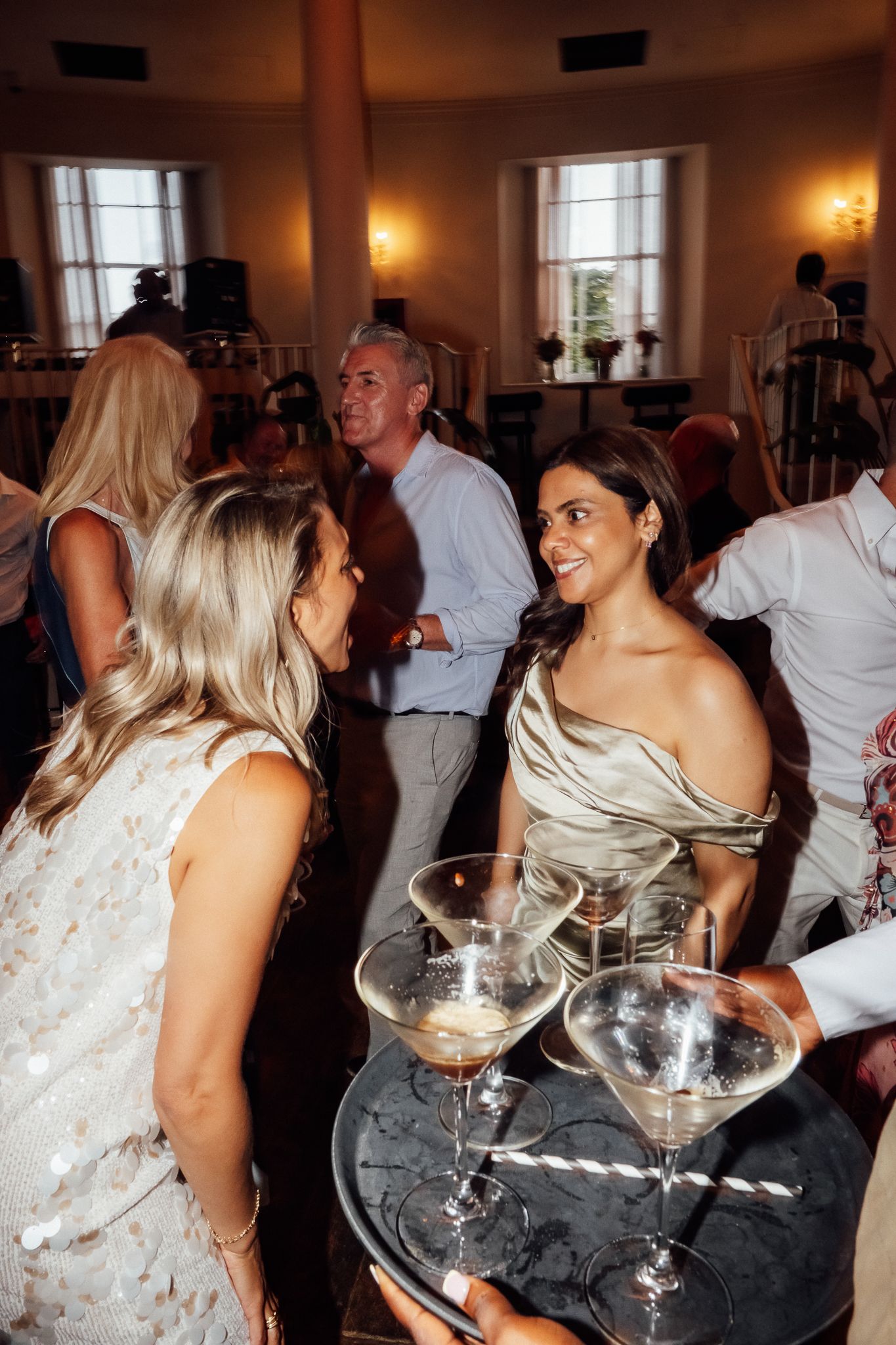 Guests at an elegant evening party smile and mingle as a tray of martini glasses is served in a warmly lit ballroom.