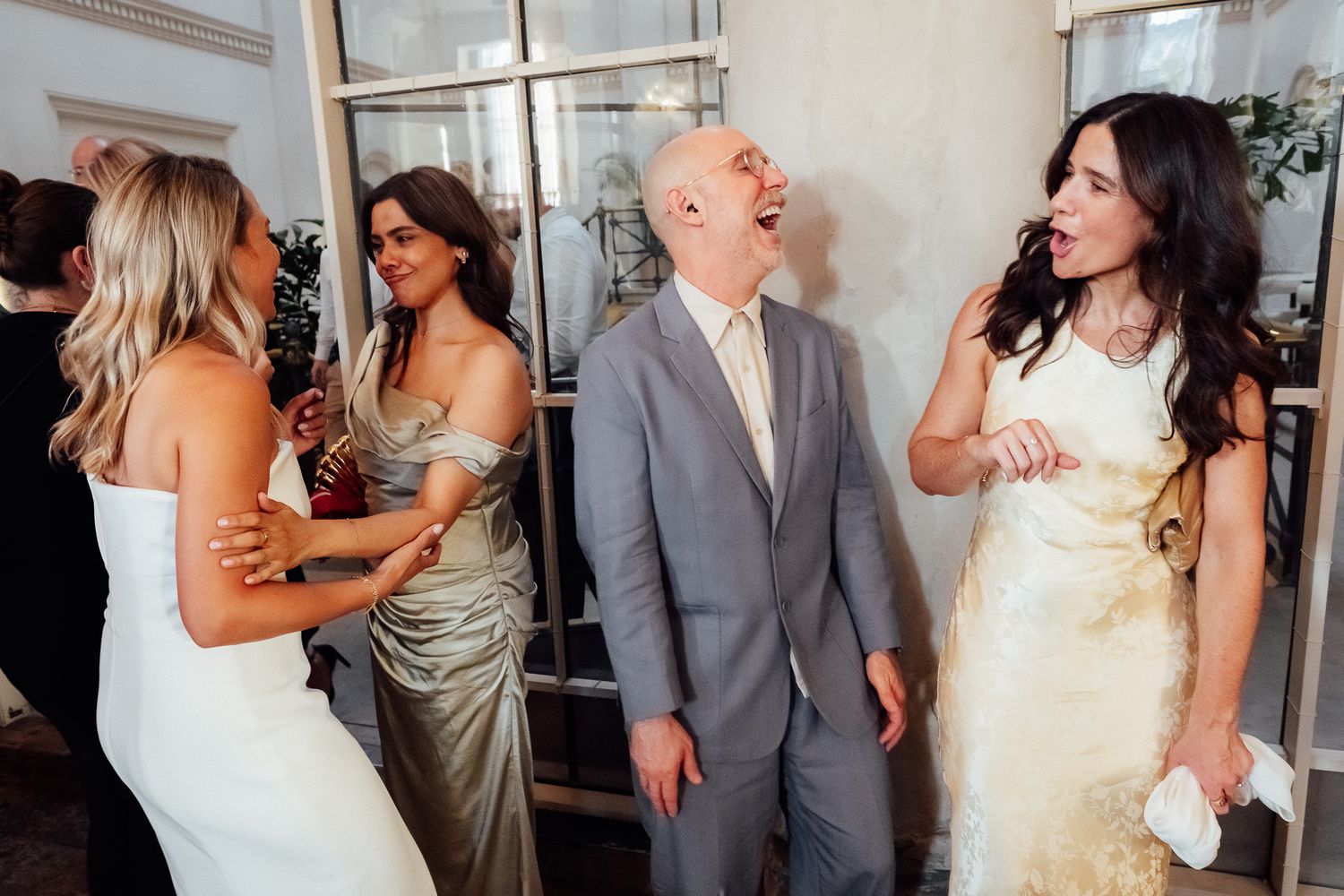 Four guests in formal attire laugh and chat at an indoor celebration, including a bald man in a gray suit and women in elegant dresses.
