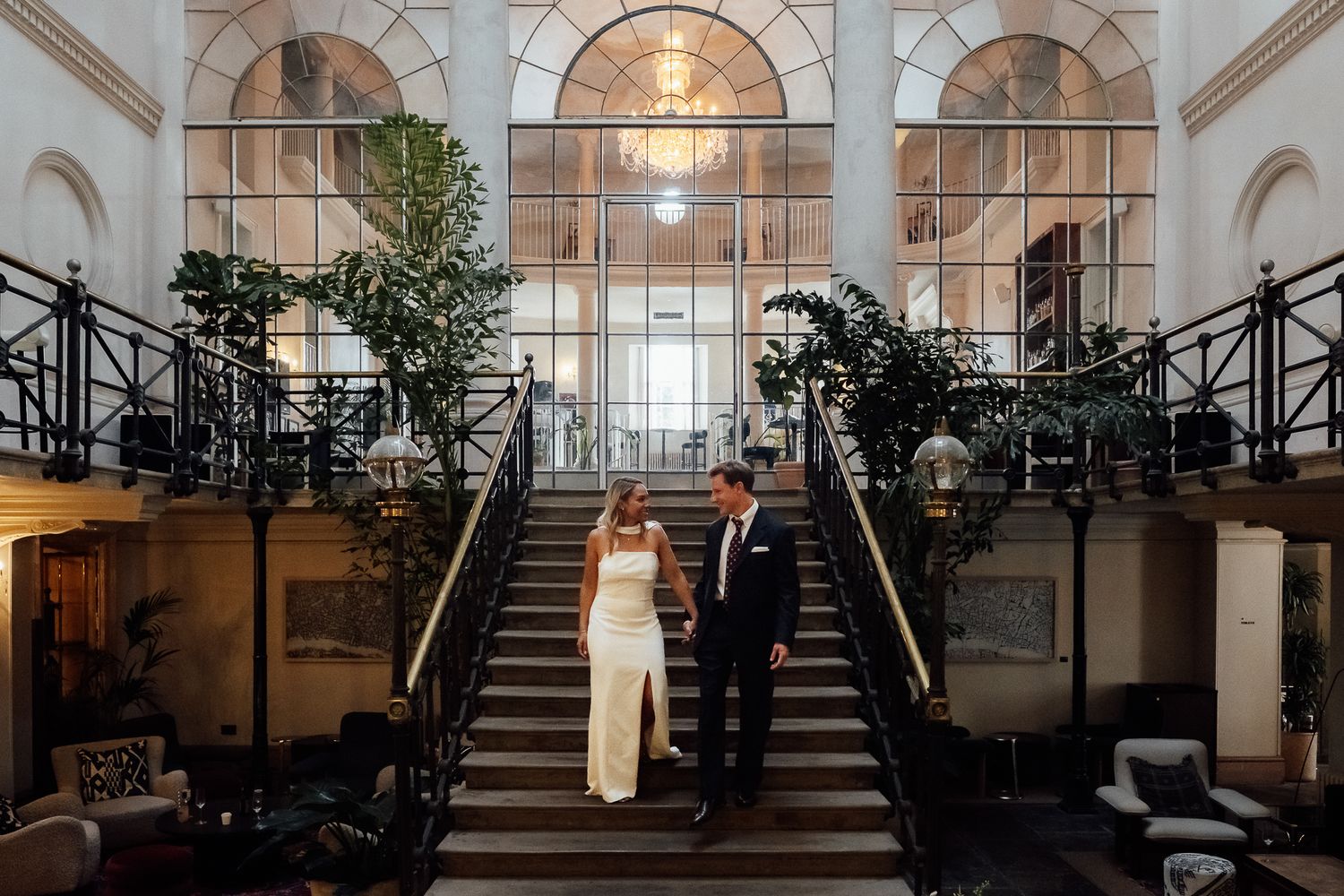 Bride in a white gown and groom in a dark suit walk down an elegant staircase in a grand venue with arched windows and lush indoor plants.