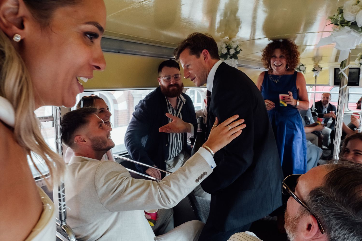 Smiling wedding guests celebrate and dance inside a decorated vintage bus as a groom in a white suit and a man in a dark suit embrace.