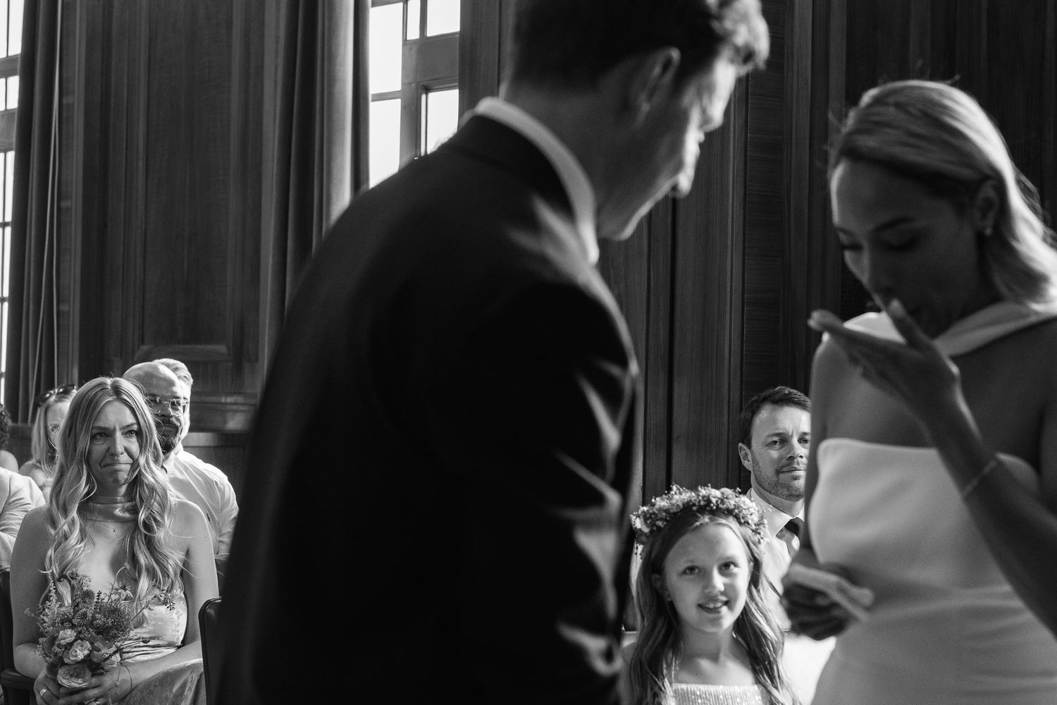 Black and white wedding ceremony photo of a bride wiping happy tears as the groom stands before her and a flower girl and guests look on.