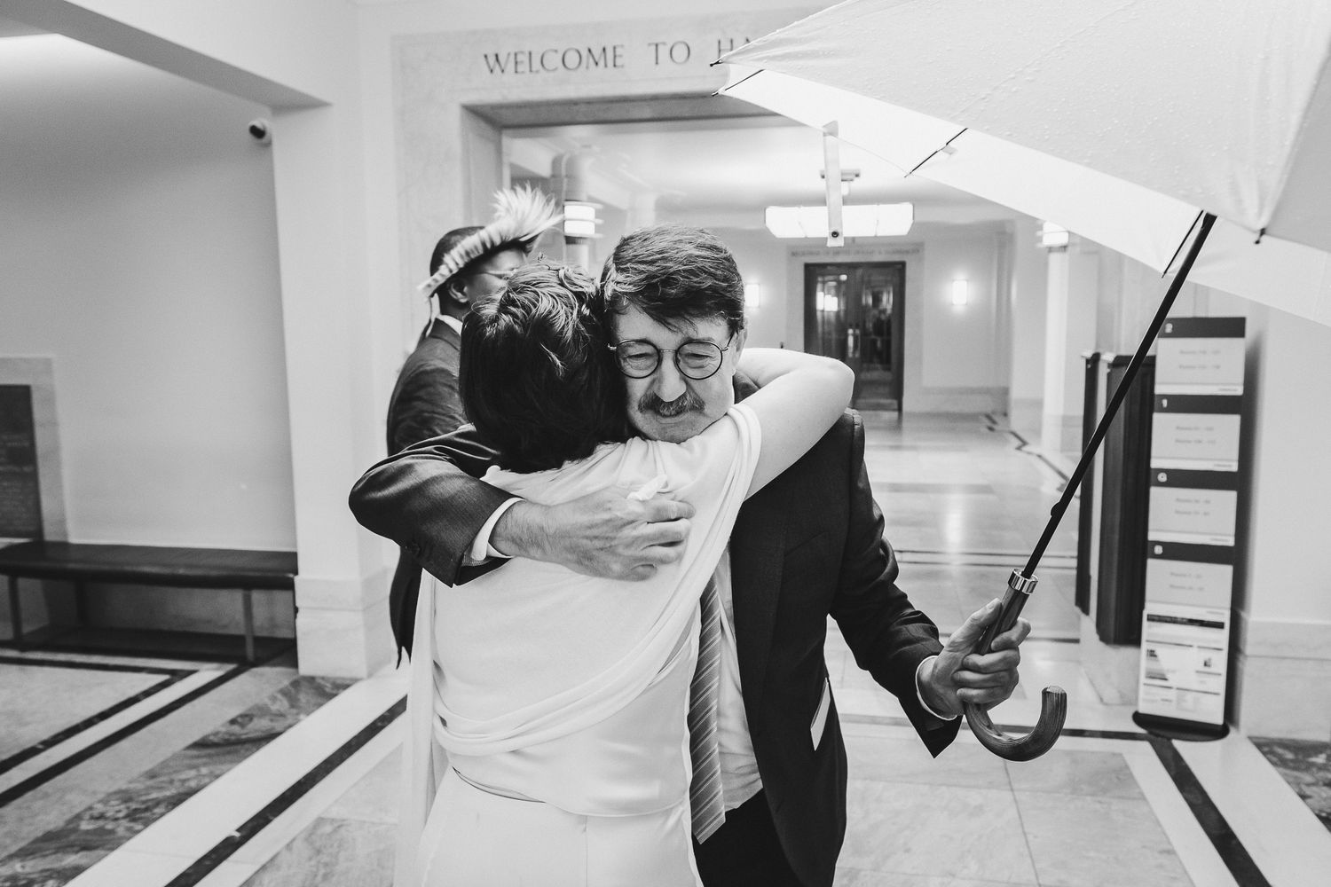 Two people embrace in a joyful moment while one holds a golf club in an indoor hallway.