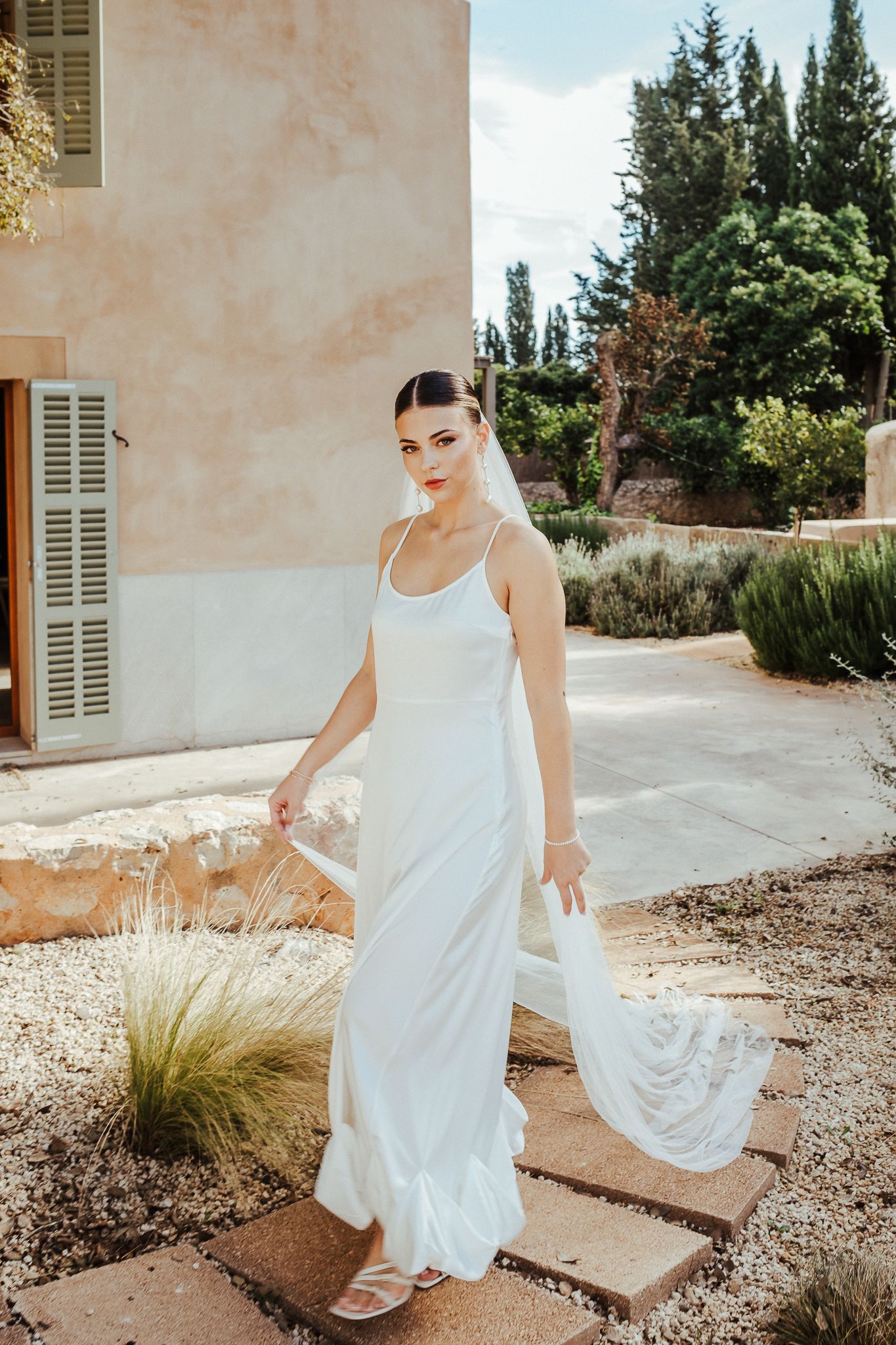 A woman in a flowing white dress twirls gracefully in a Mediterranean garden setting with rustic walls and ornamental grasses.