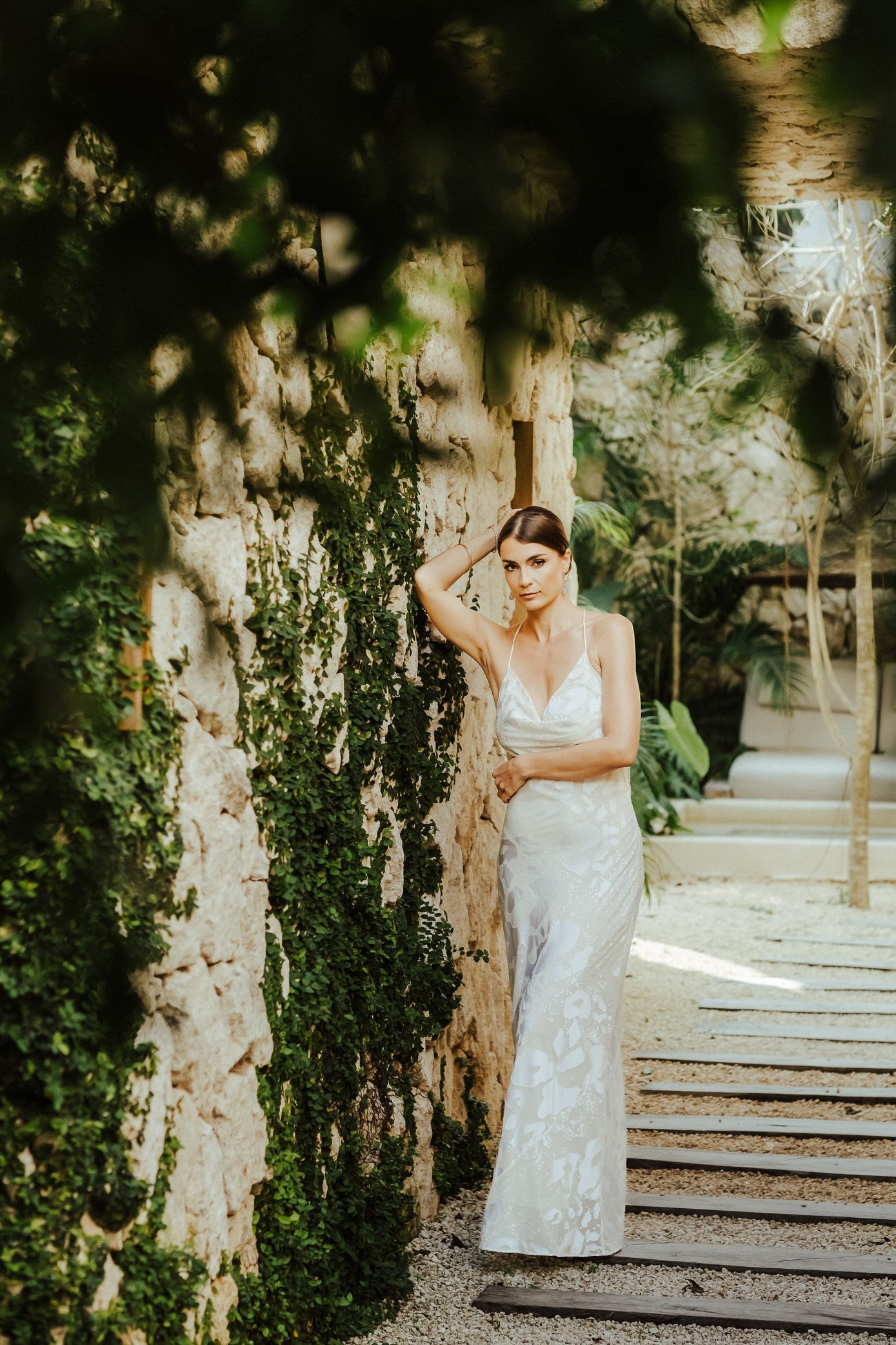 A person in a white lace wedding dress poses gracefully among trees in a garden setting.