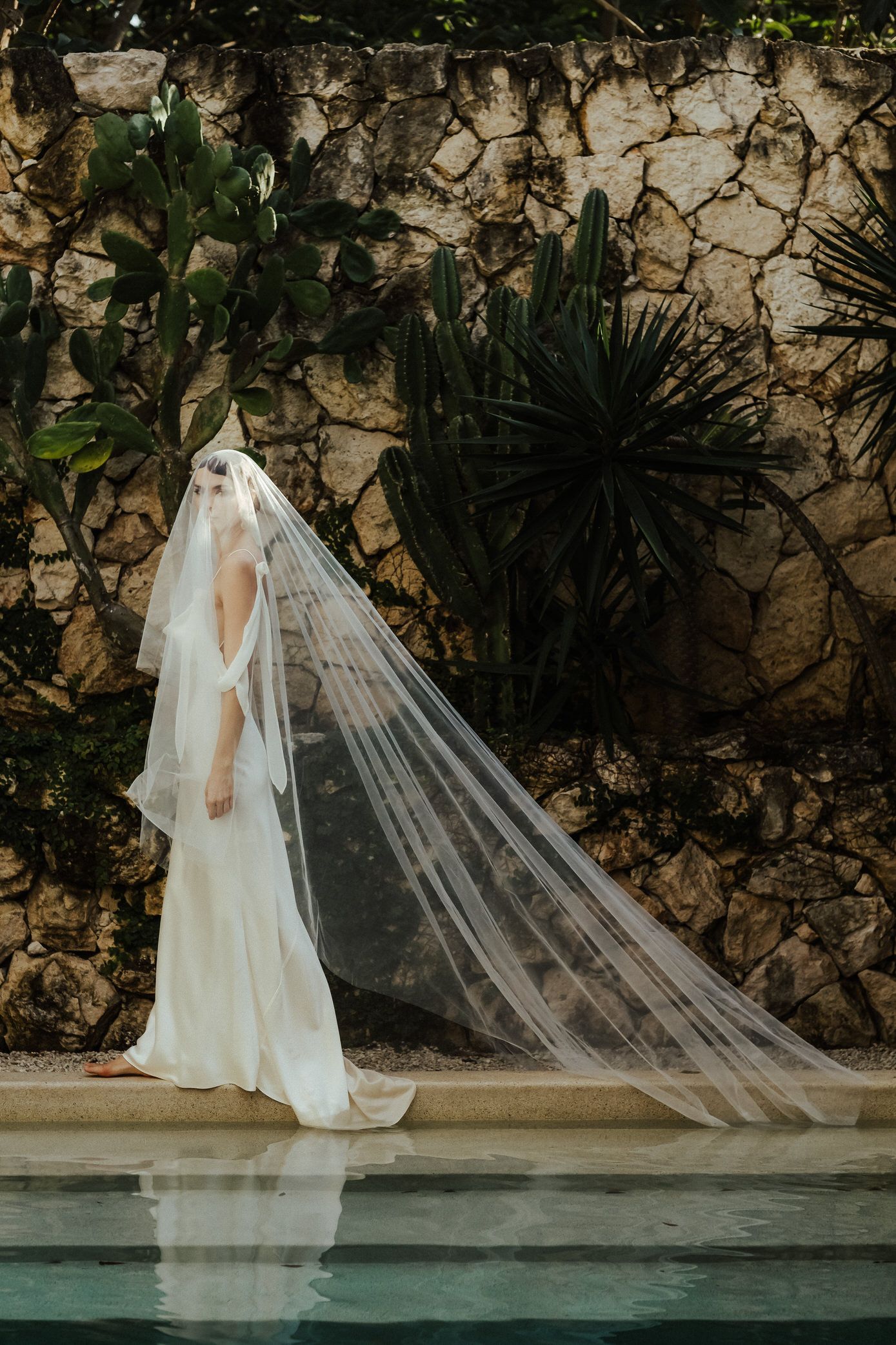 A bride in a flowing white wedding dress and long veil walks elegantly along a poolside stone wall.