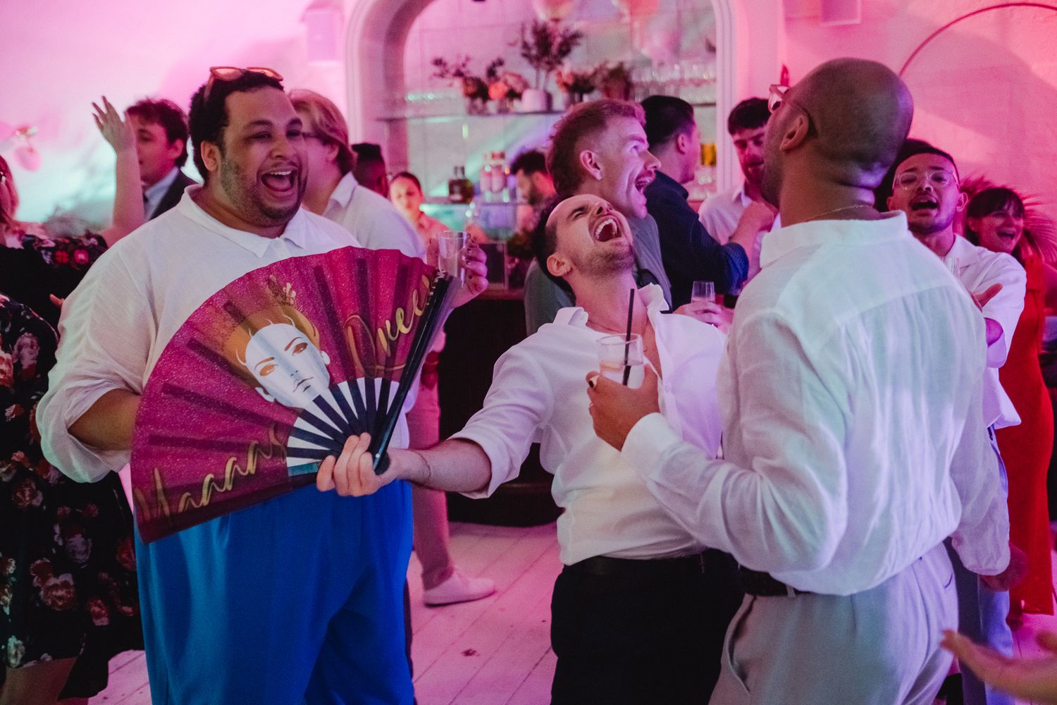 Party guests dance and celebrate with pink lighting and decorative fans at an indoor event with festive ambiance.