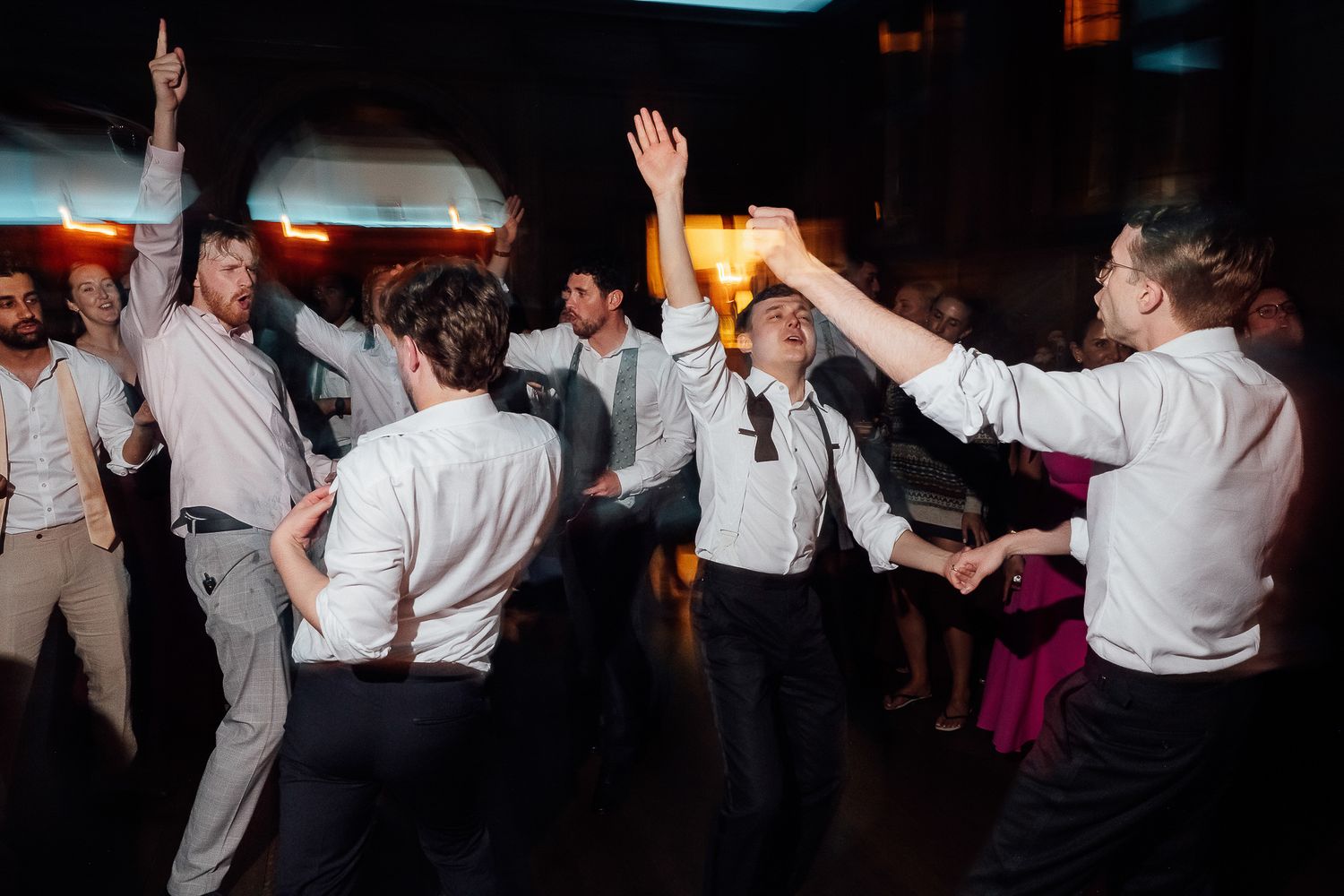 Men in white dress shirts dancing energetically at a wedding reception party on a dark dance floor.