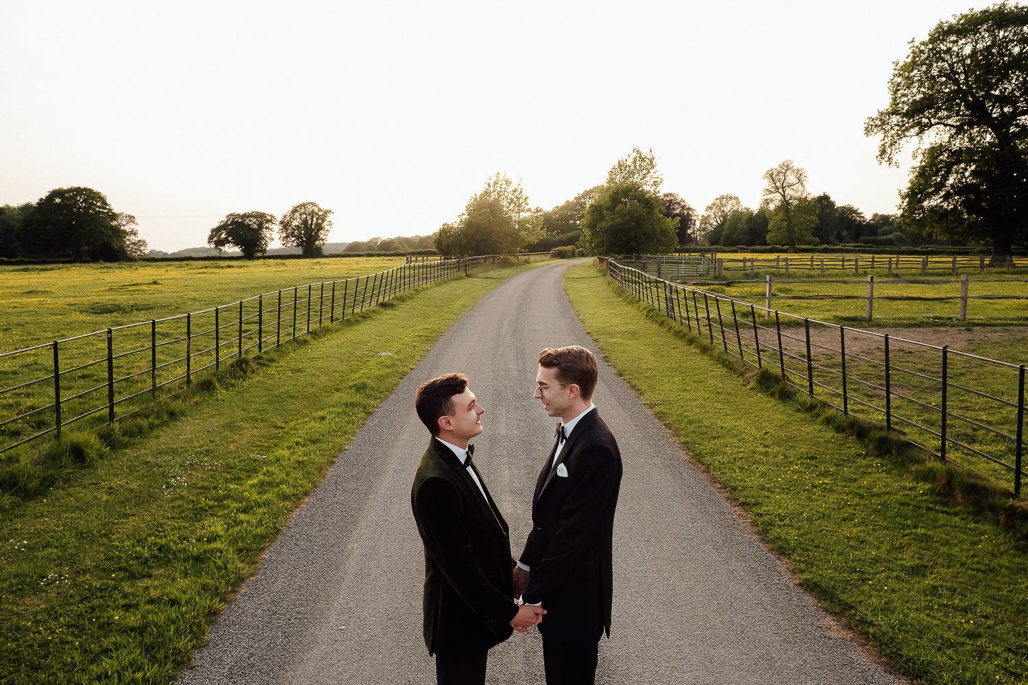 Two grooms in black tuxedos hold hands on a scenic gravel path at sunset during their outdoor wedding ceremony.