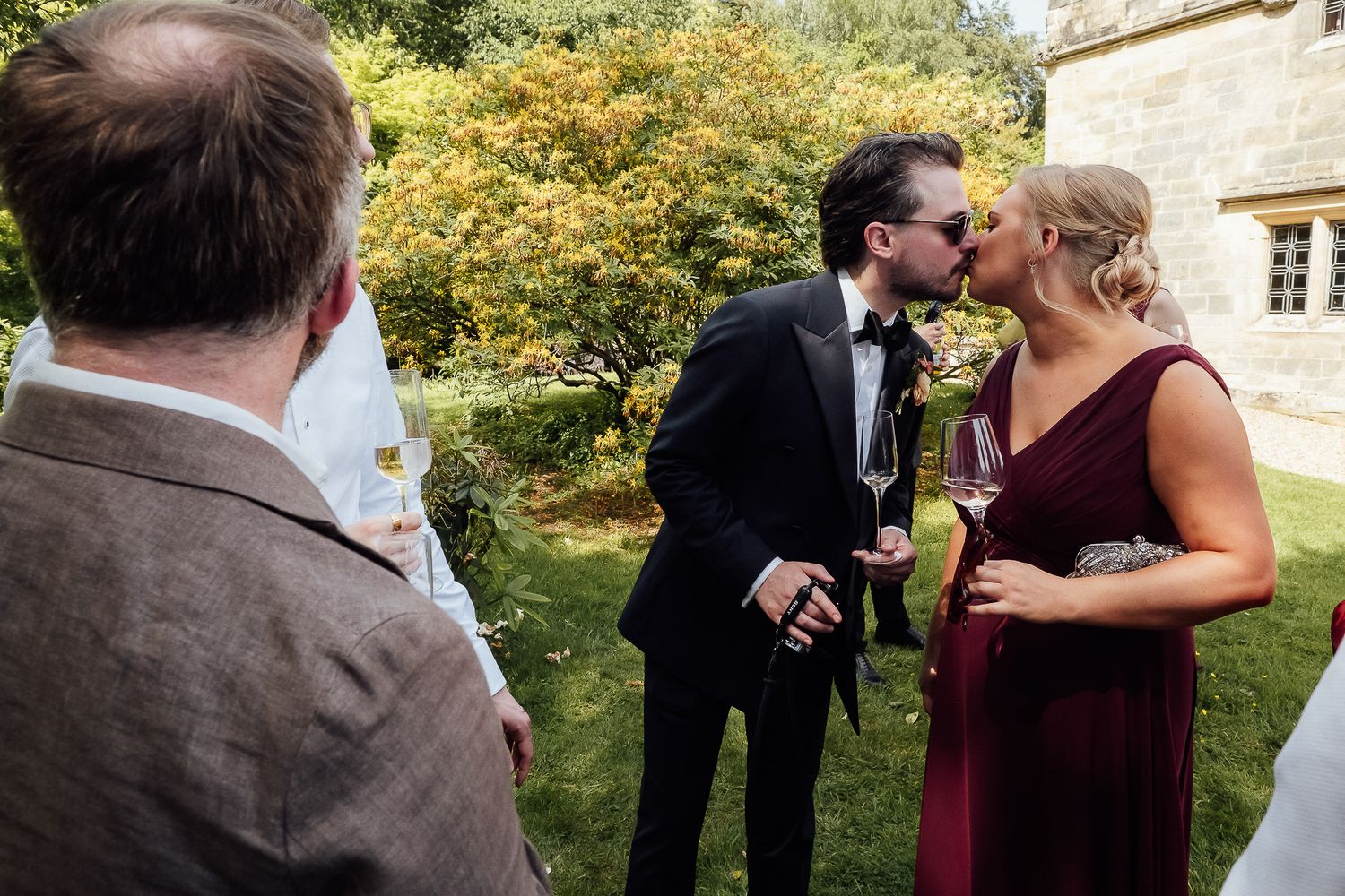 Couple sharing a kiss at outdoor wedding reception, groom in tuxedo and bride in burgundy dress holding champagne glasses.