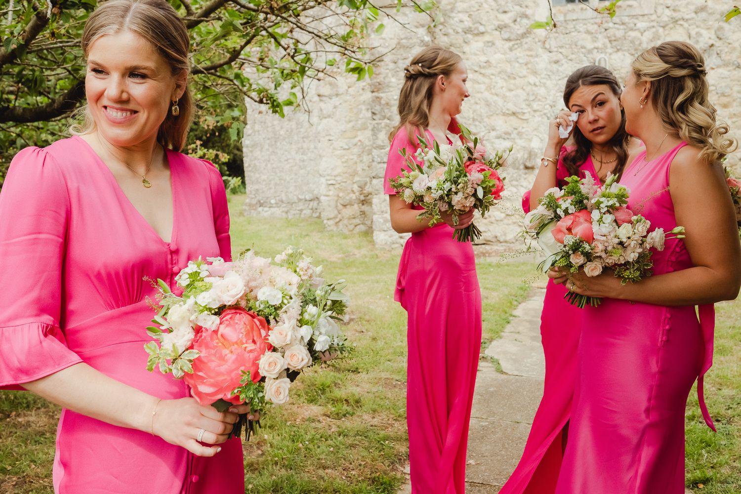 Bridesmaids in bright pink dresses gather in an outdoor garden setting holding flower bouquets.