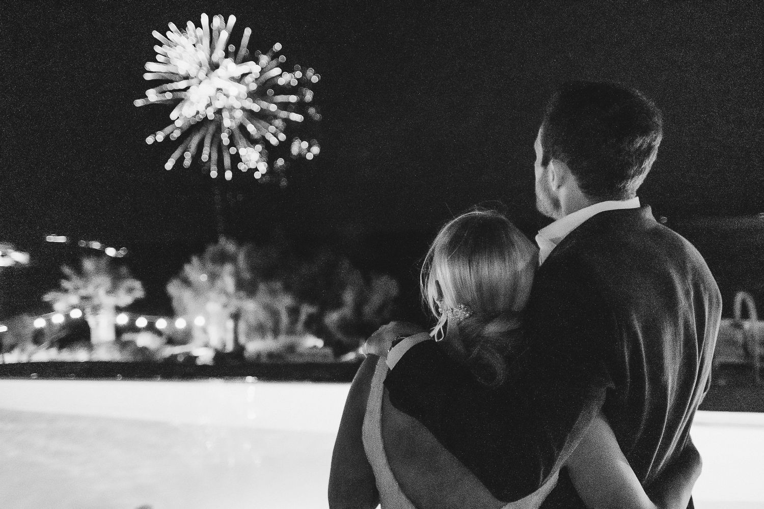 Silhouetted couple embraces against a sunset backdrop with fireworks bursting in the sky above them.