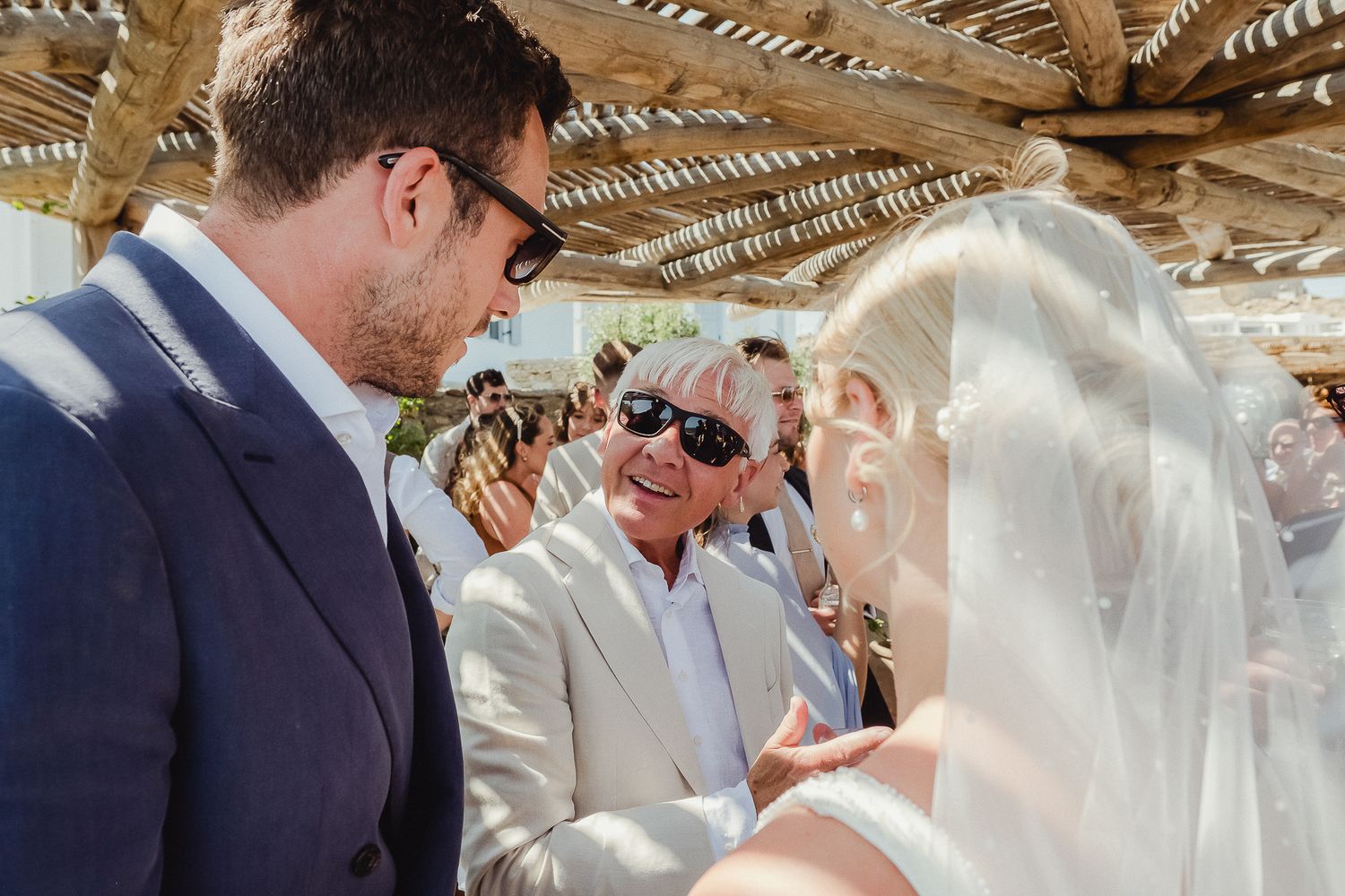 A beach wedding ceremony takes place under a shaded canopy with guests watching the couple exchange vows at sunset.
