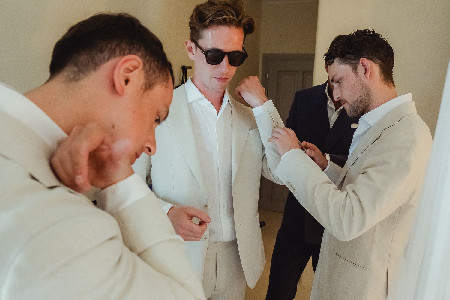 Six men in white suits and one in a black suit stand in a line while holding their faces in humorous poses.