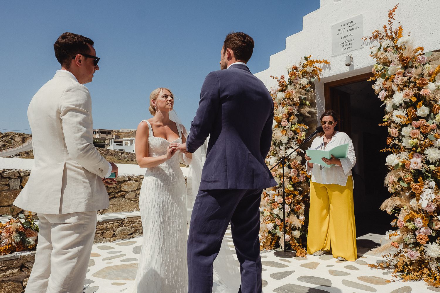 Outdoor wedding ceremony under a floral arch with peach and cream flowers on a whitewashed building in Greece.