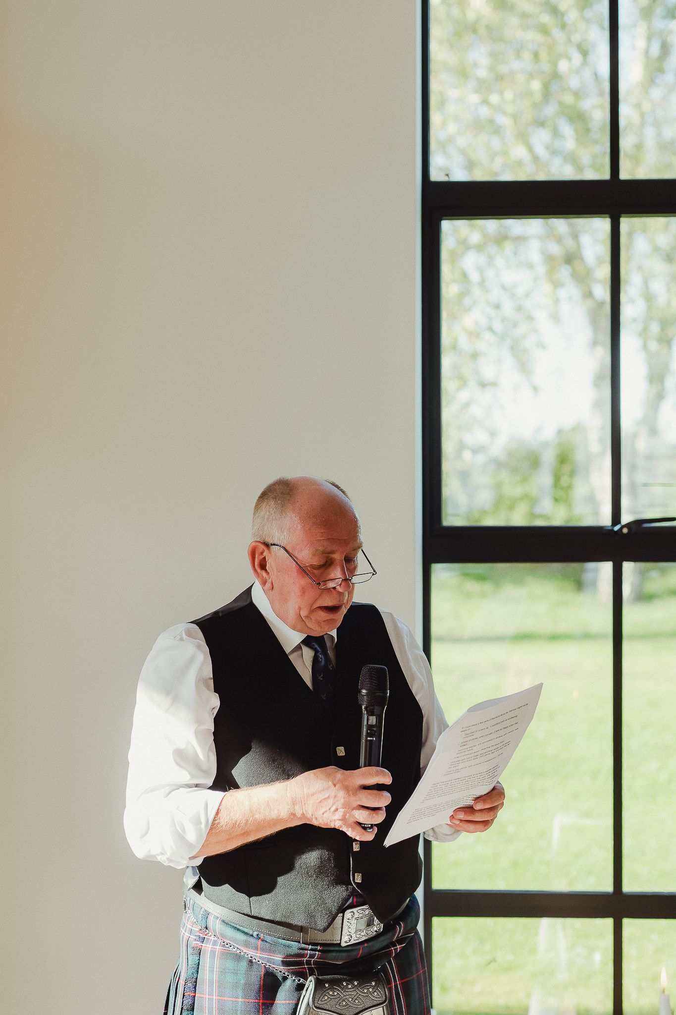 A person in a vest and kilt reads documents near large windows in a bright interior space.