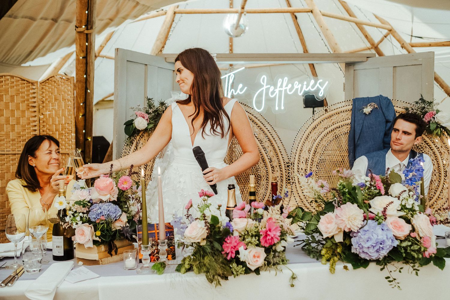 Wedding reception guests share laughter at a table decorated with elegant floral arrangements and neon signage.