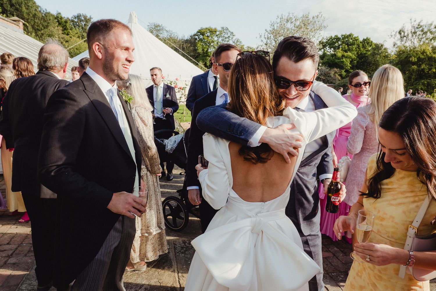 Wedding guests gather outdoors during a reception as a couple embraces in a backless white dress and formal attire.