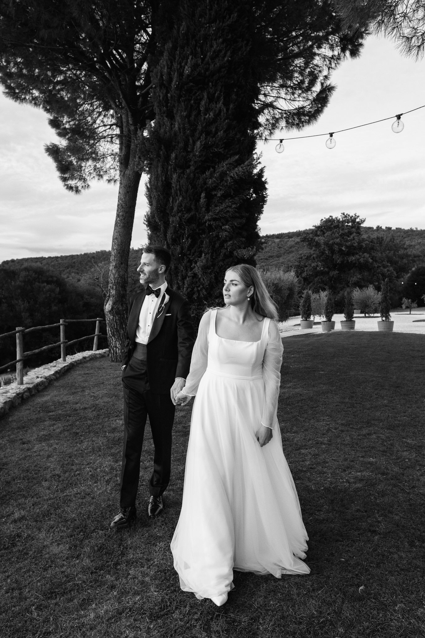 Black and white photo of a bride and groom holding hands on a lawn beneath a tall cypress tree, with string lights and rolling hills behind.