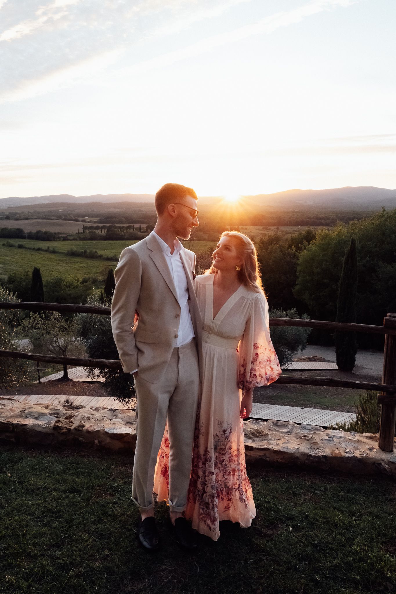 Couple in beige suit and floral maxi dress embrace at sunset on a stone terrace overlooking rolling Tuscan hills.