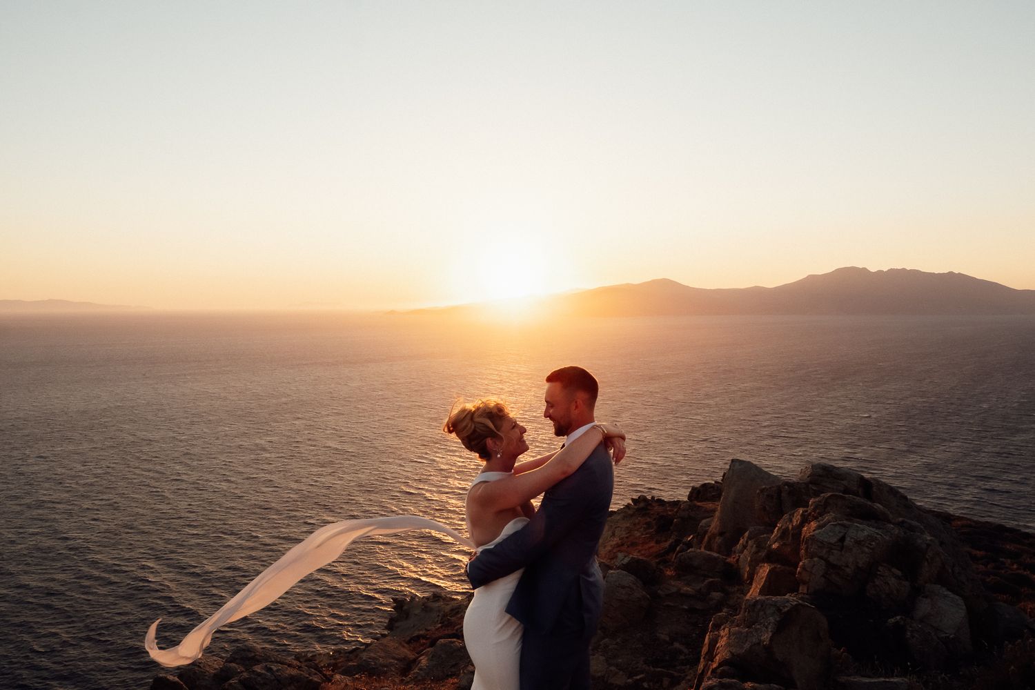 Bride and groom embrace on rocky cliffside at golden sunset over ocean, wedding veil flowing in breeze.