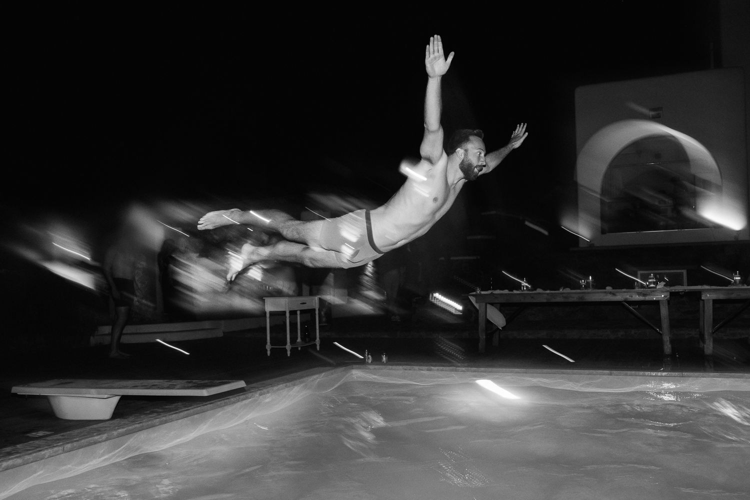 Black and white photo of a man diving off a springboard into a pool at night, captured with motion blur.