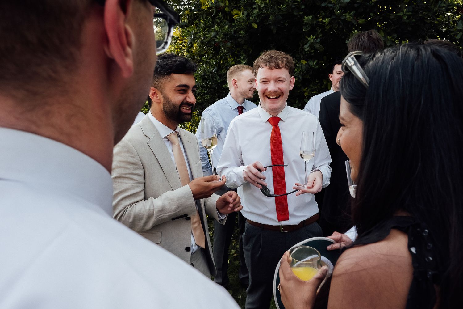 Group of guests laughing and socializing at an outdoor wedding reception, man in red tie smiling broadly.