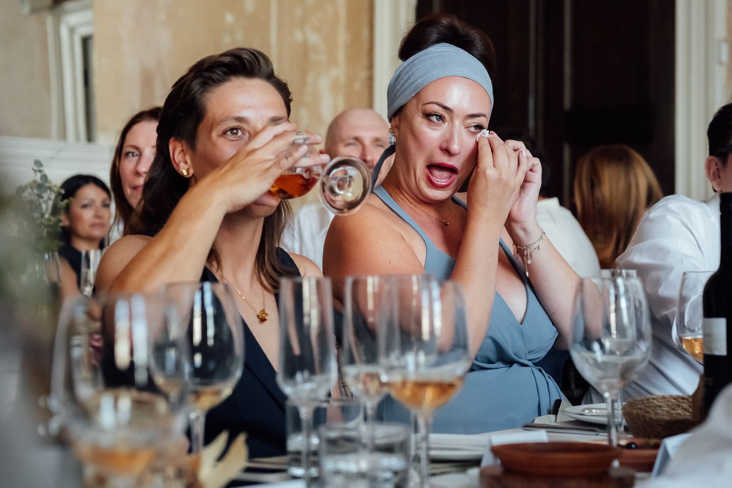 Two women at a crowded banquet table laugh and sip drinks, one in a blue headband wiping her eye, with wine glasses in the foreground.