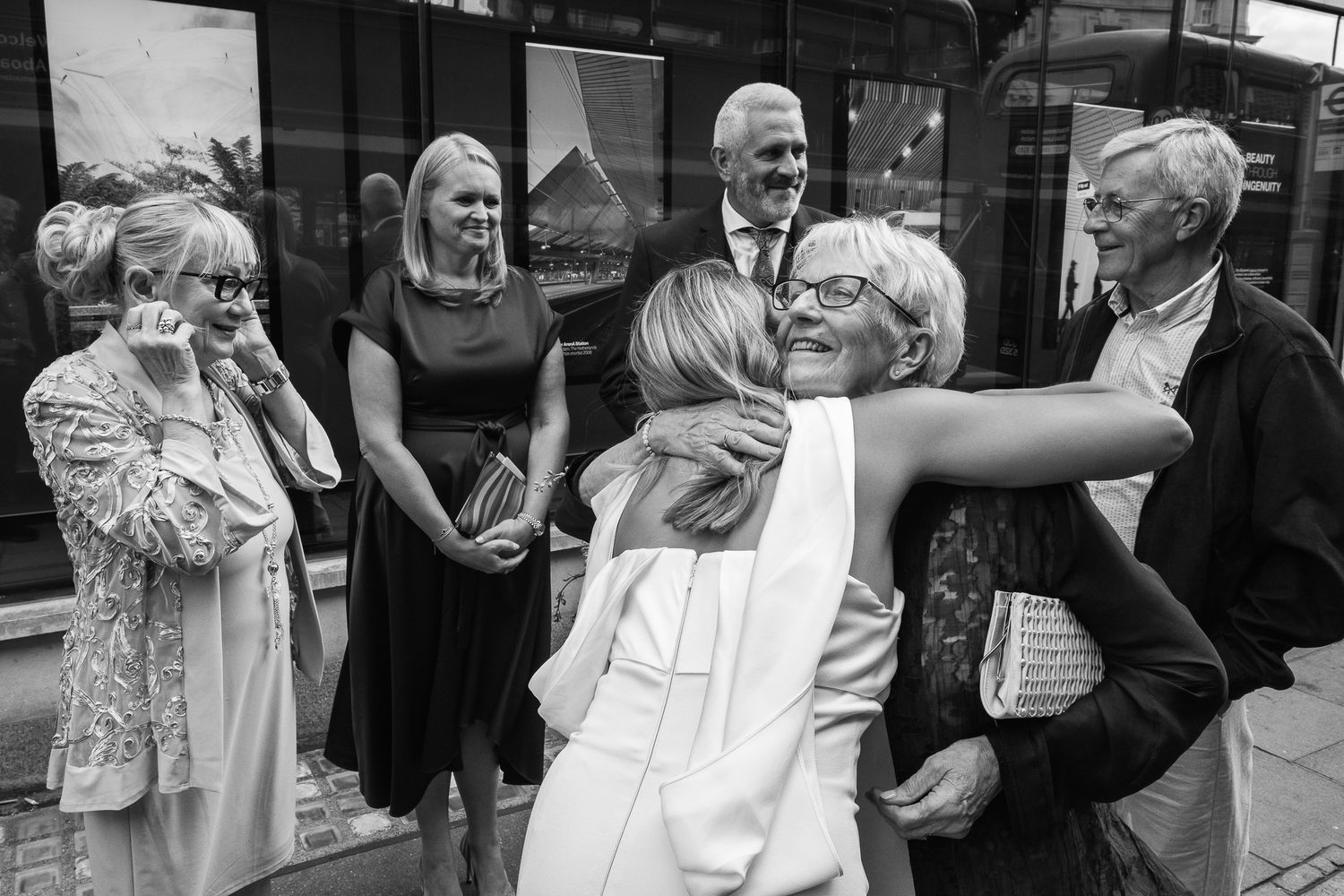 Black and white photo of two women embracing warmly while smiling friends and family look on outside a building.
