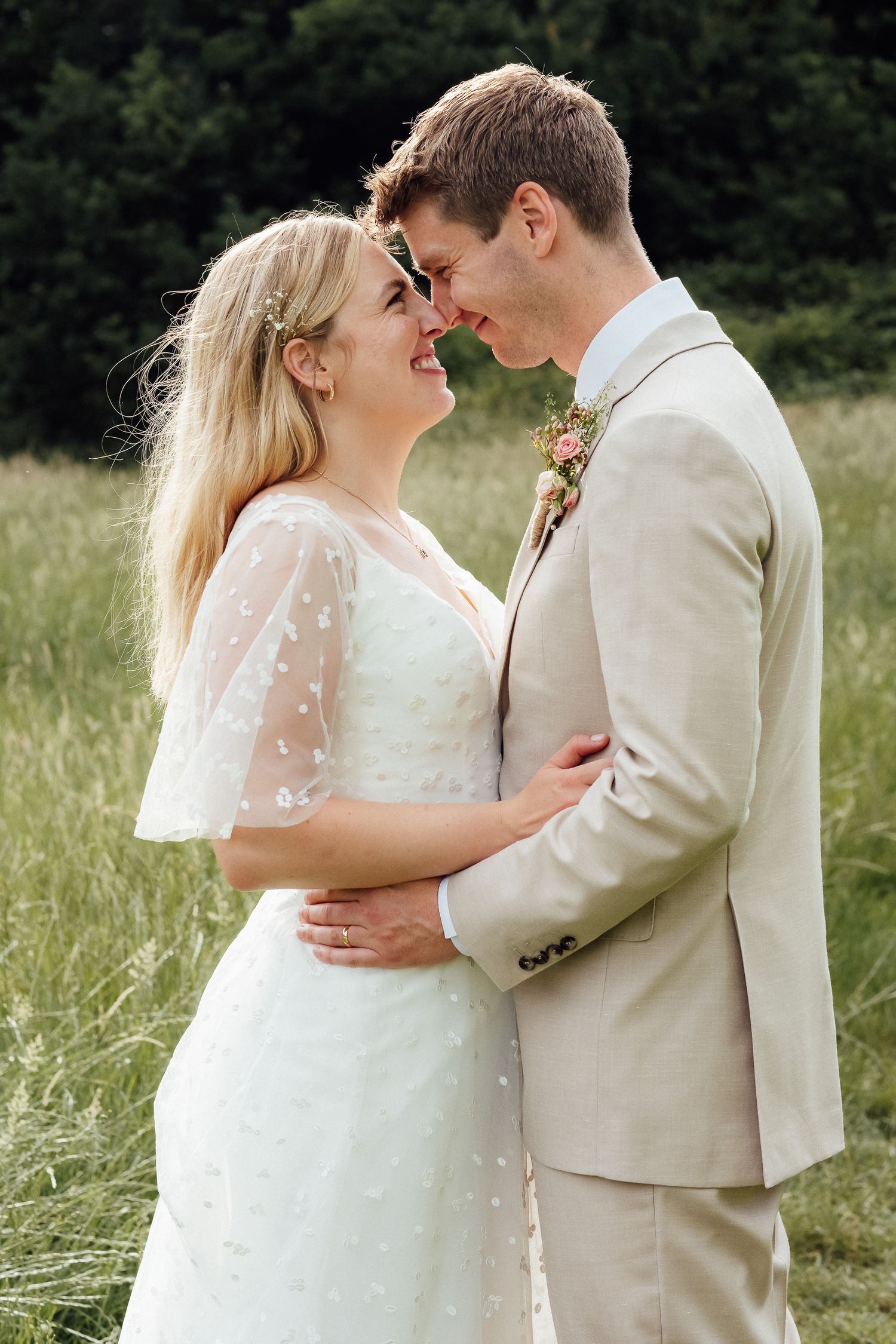 Smiling bride in a white lace dress and groom in a beige suit with a floral boutonniere embrace nose to nose in a tall grass meadow.