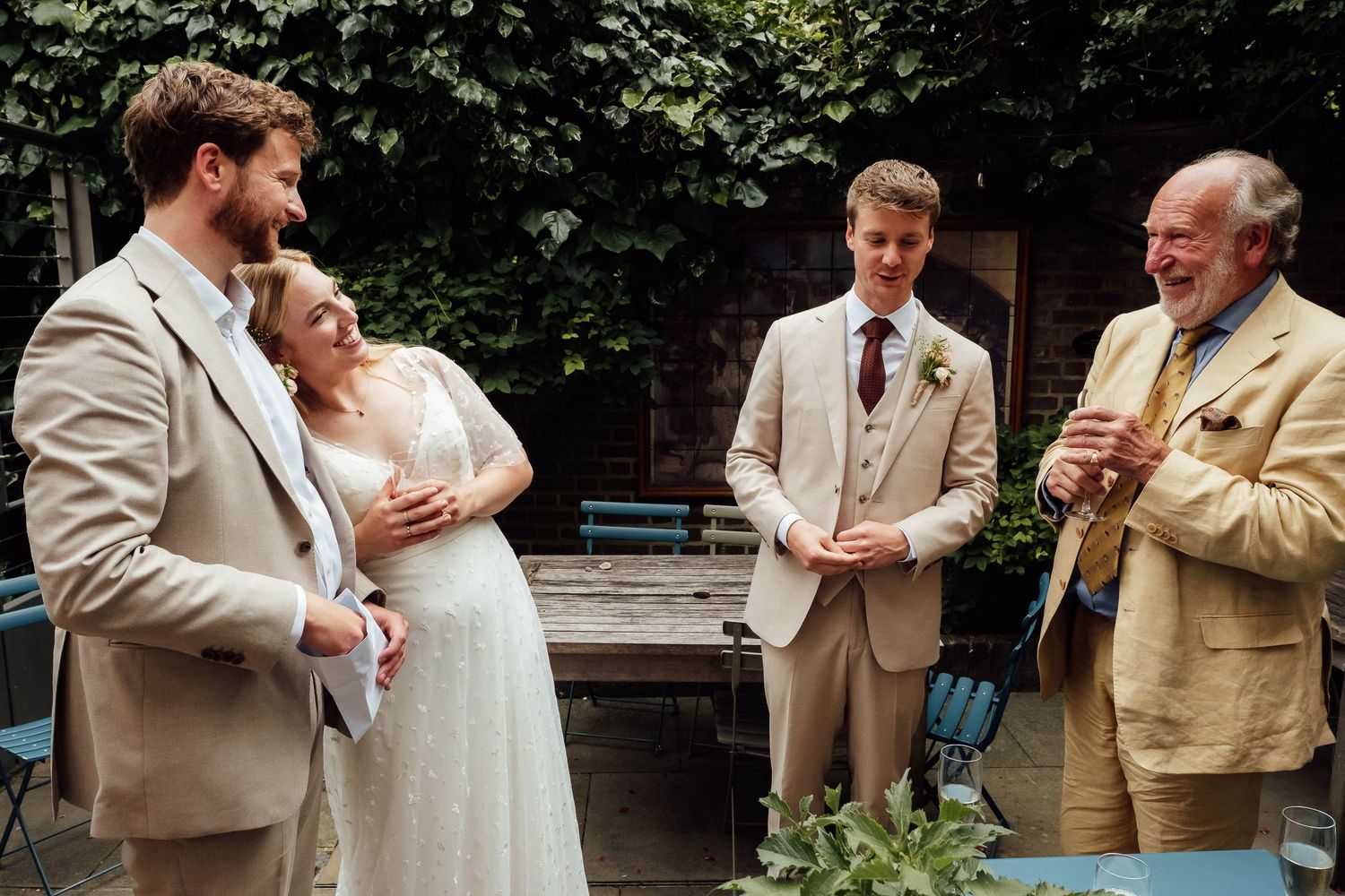 A bride in a white gown laughs with a bearded man as a groom and an older man in tan suits chat in a leafy garden courtyard.