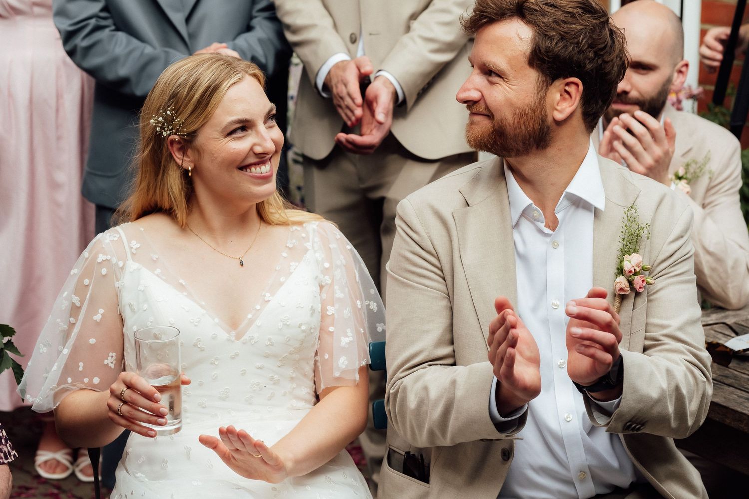 Smiling bride in a white lace gown and a bearded groom in a tan suit applaud joyfully while wedding guests clap behind them.