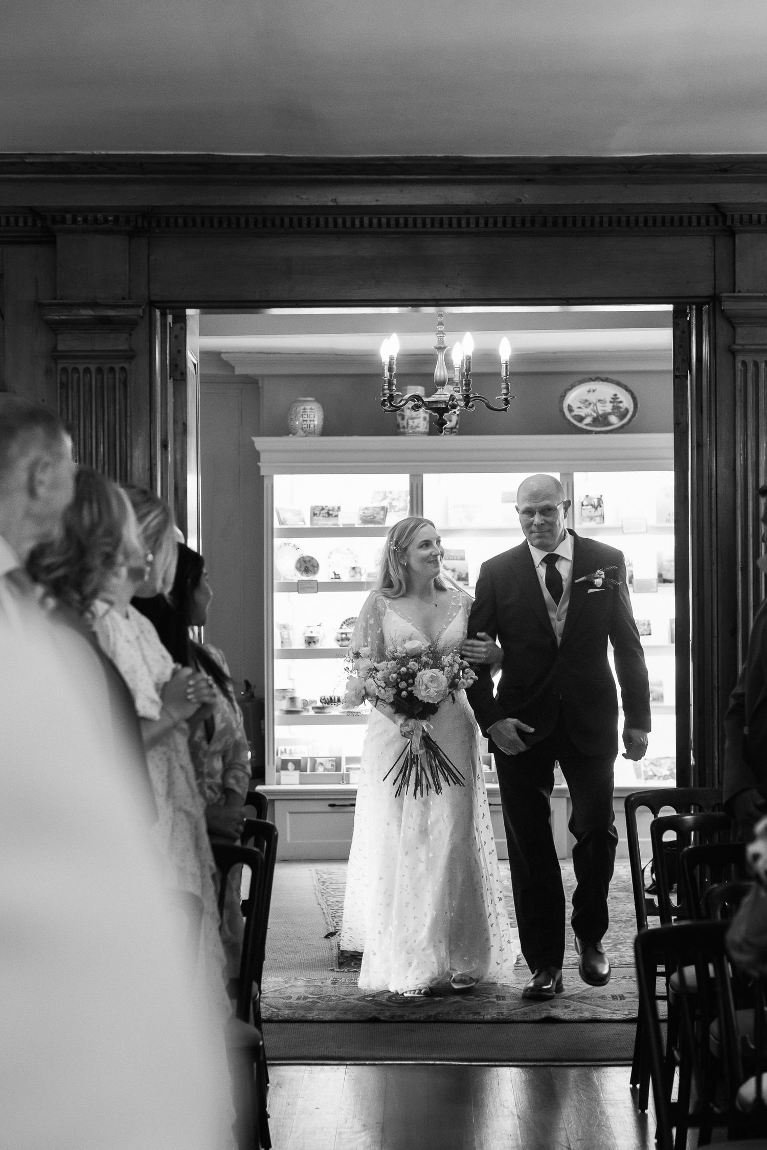 A father in a dark suit walks his bride in a white lace gown carrying a bouquet down the aisle in a black and white photo.