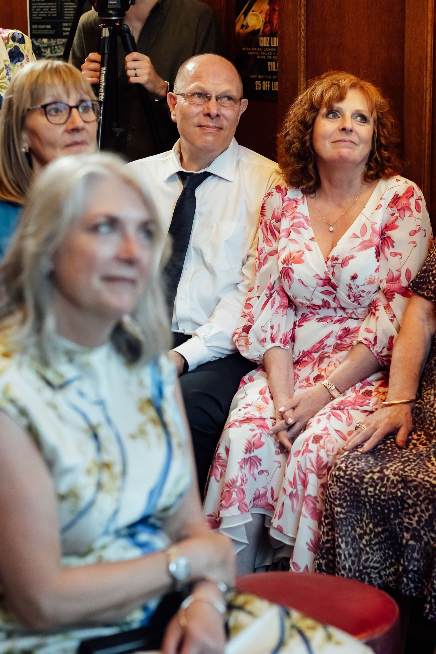 Group of smiling adults seated closely together in an audience, including a bald man in a tie and women in floral and patterned dresses.