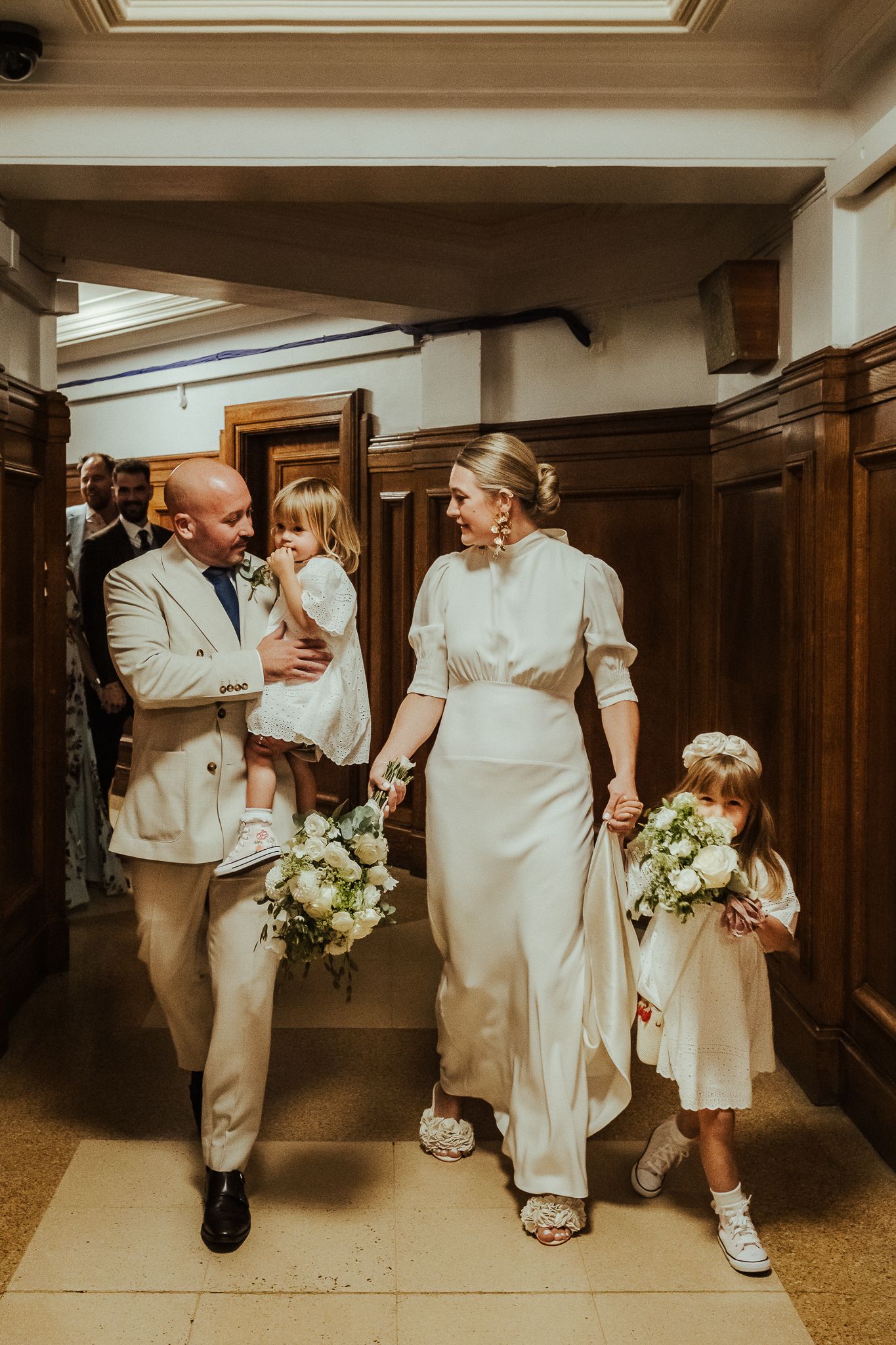 A group of people in white dresses and tan suits walk together down a wooden-paneled hallway at a wedding celebration.