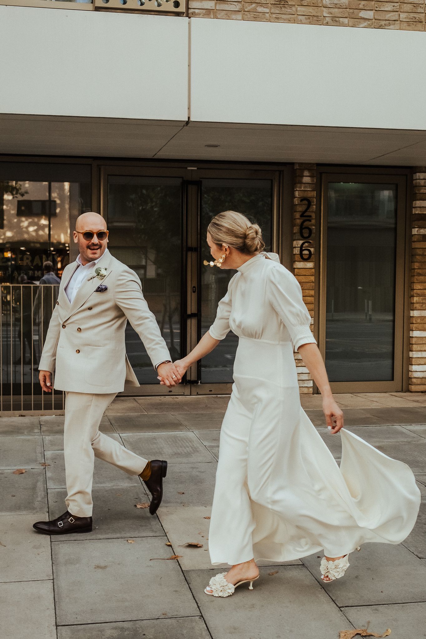 A couple wearing matching cream-colored wedding attire hold hands and walk together on a downtown sidewalk.