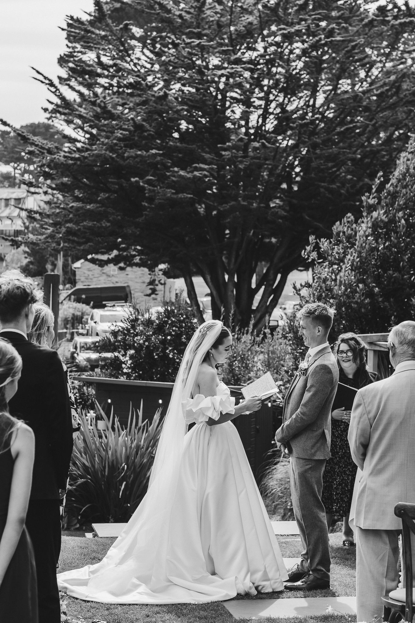 Wedding party gathers outdoors under cypress trees during a ceremony by the ocean.
