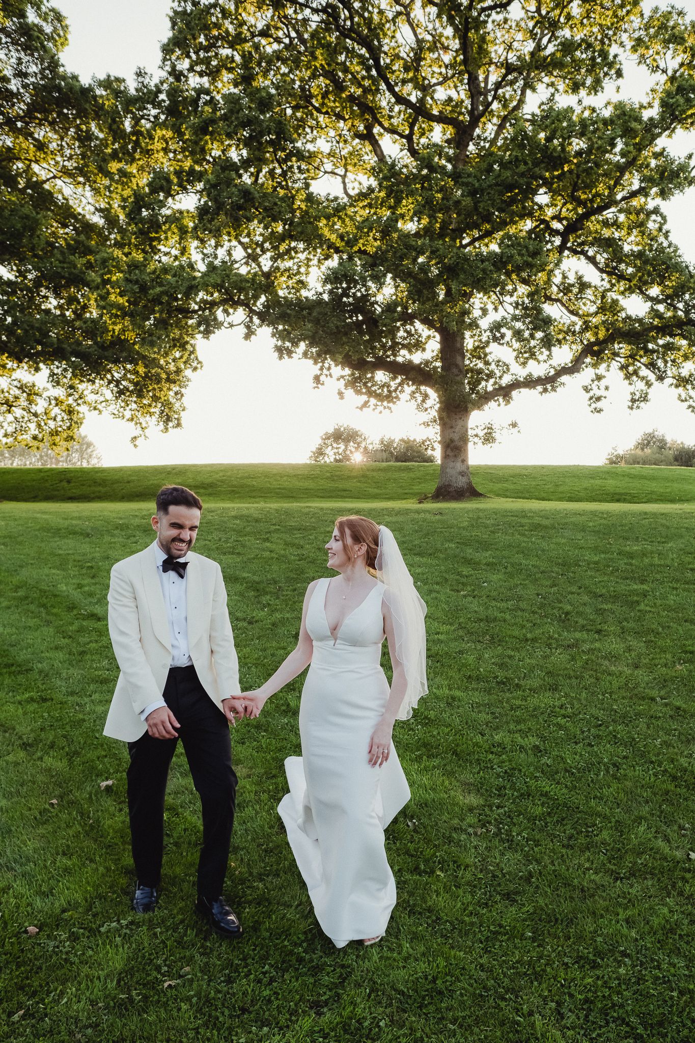 A couple shares a joyful moment together on a grassy hill beneath a large oak tree during their outdoor wedding session.