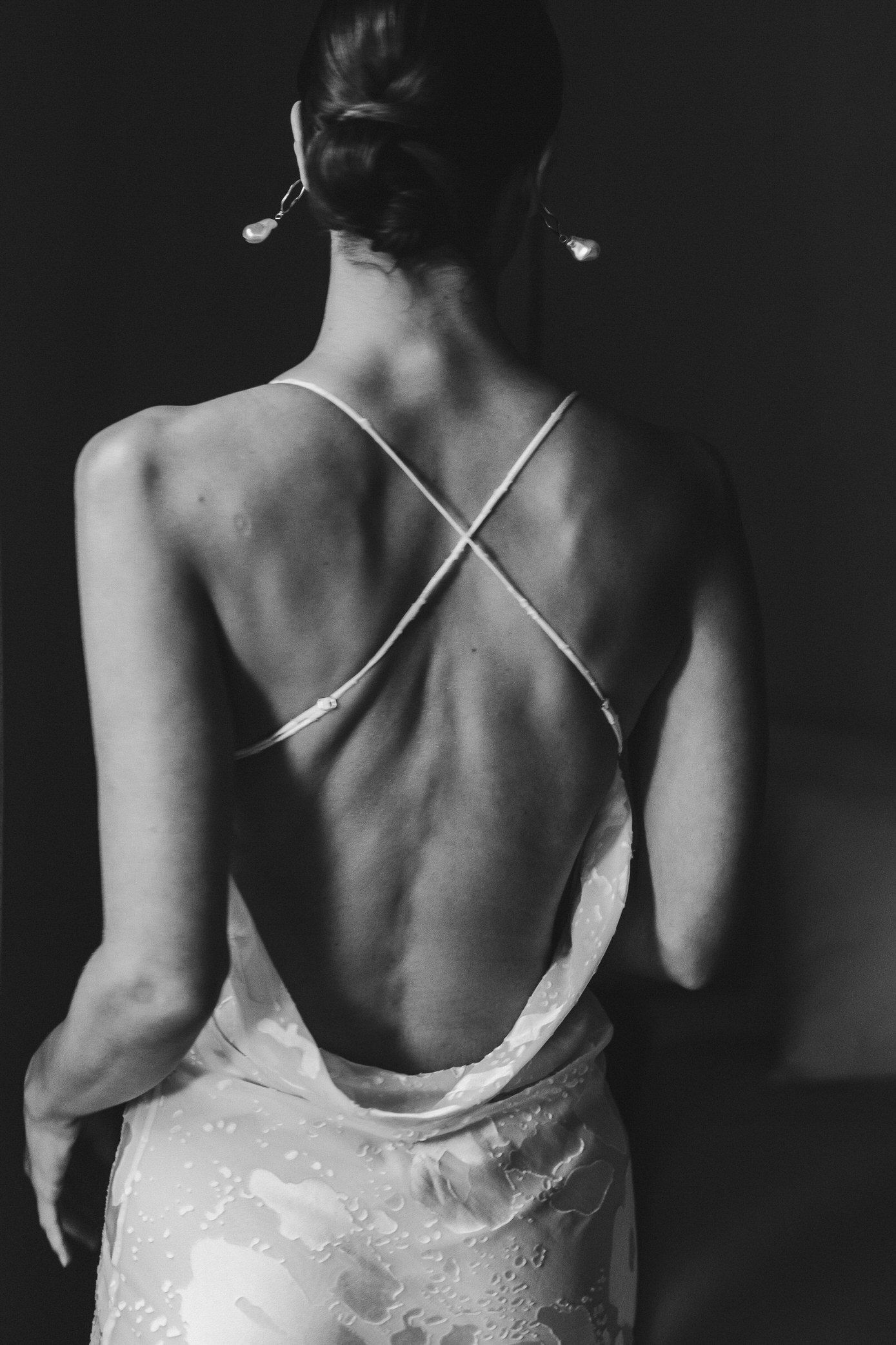Black and white photograph showing the back of a person wearing a delicate white lace dress with thin straps.