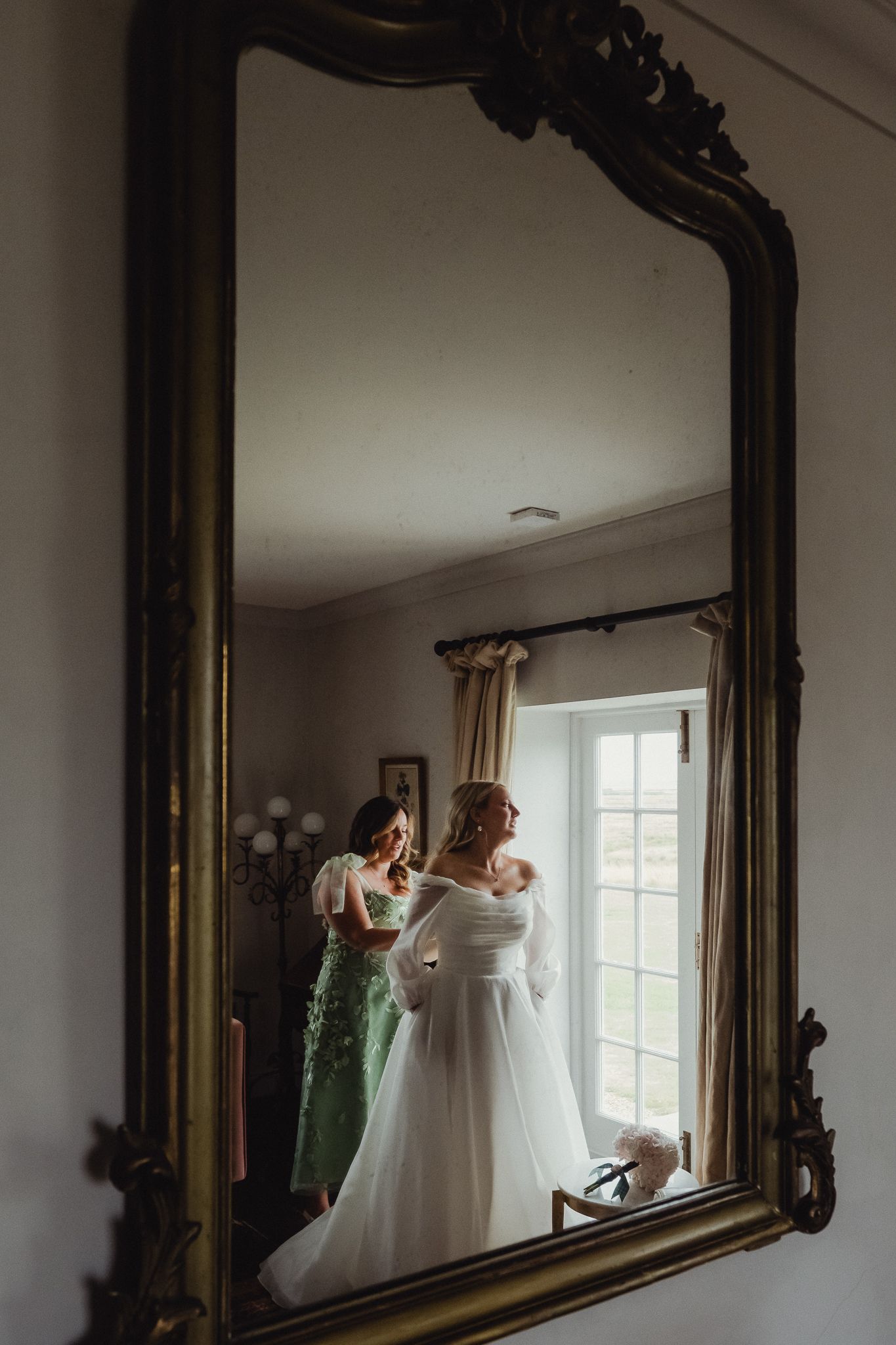 Ornate gold mirror reflects wedding preparations with dresses hanging in a sunlit room.