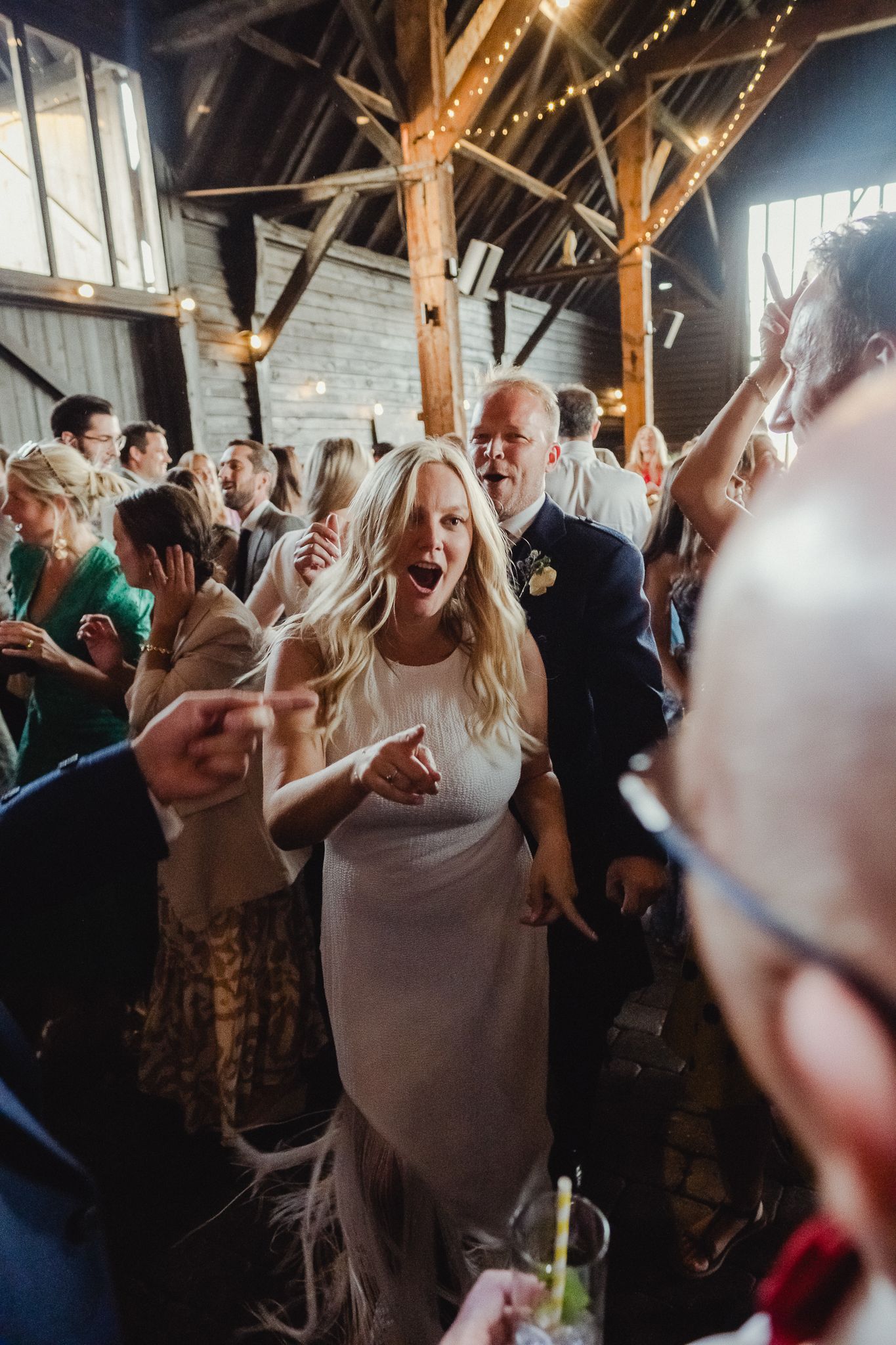 Wedding guests dance and celebrate energetically on a crowded dance floor under wooden beams at an indoor reception.