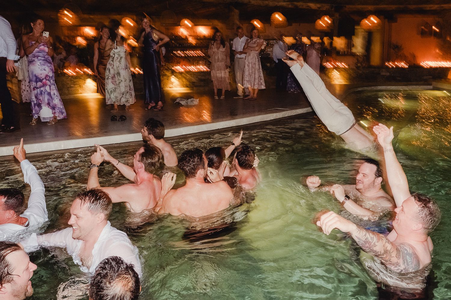 Wedding guests celebrate in a swimming pool at night with mood lighting during a festive reception party.